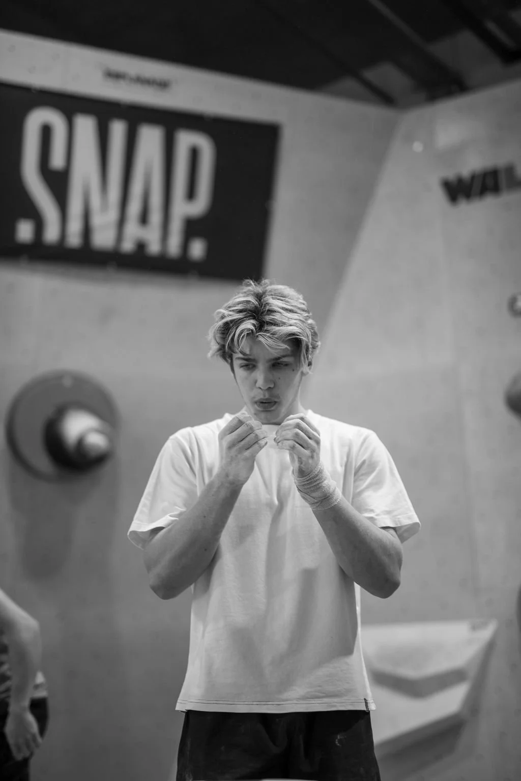 A young man with tousled hair and a white t-shirt is inside a gym, looking down and inspecting his hands. There is a sign that says 'SNAP' visible in the background, along with gym equipment and a large wall.