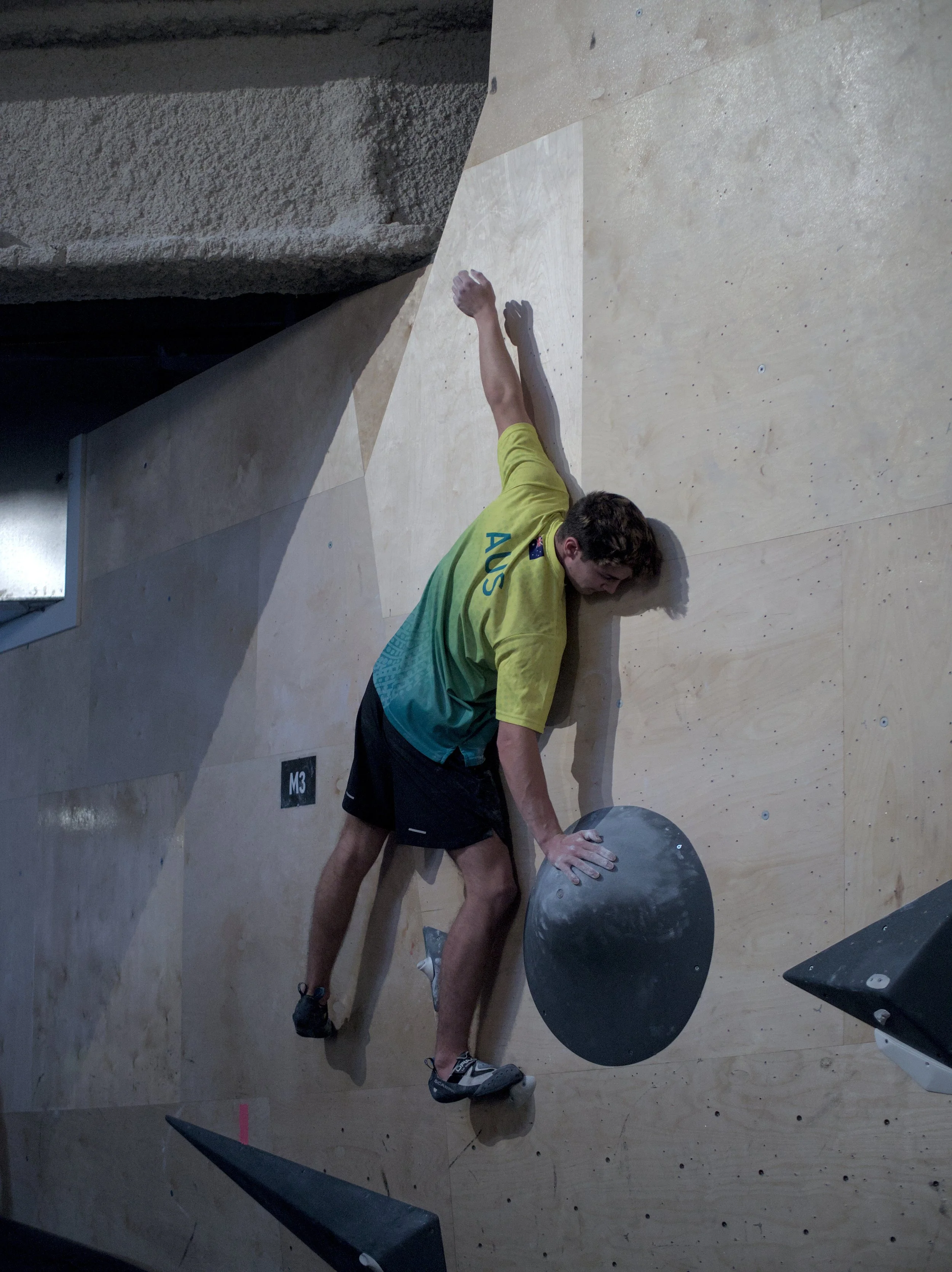 Male climber resting on climbing wall holding an orange chalk bag, wearing a yellow and green shirt, black shorts, and climbing shoes.