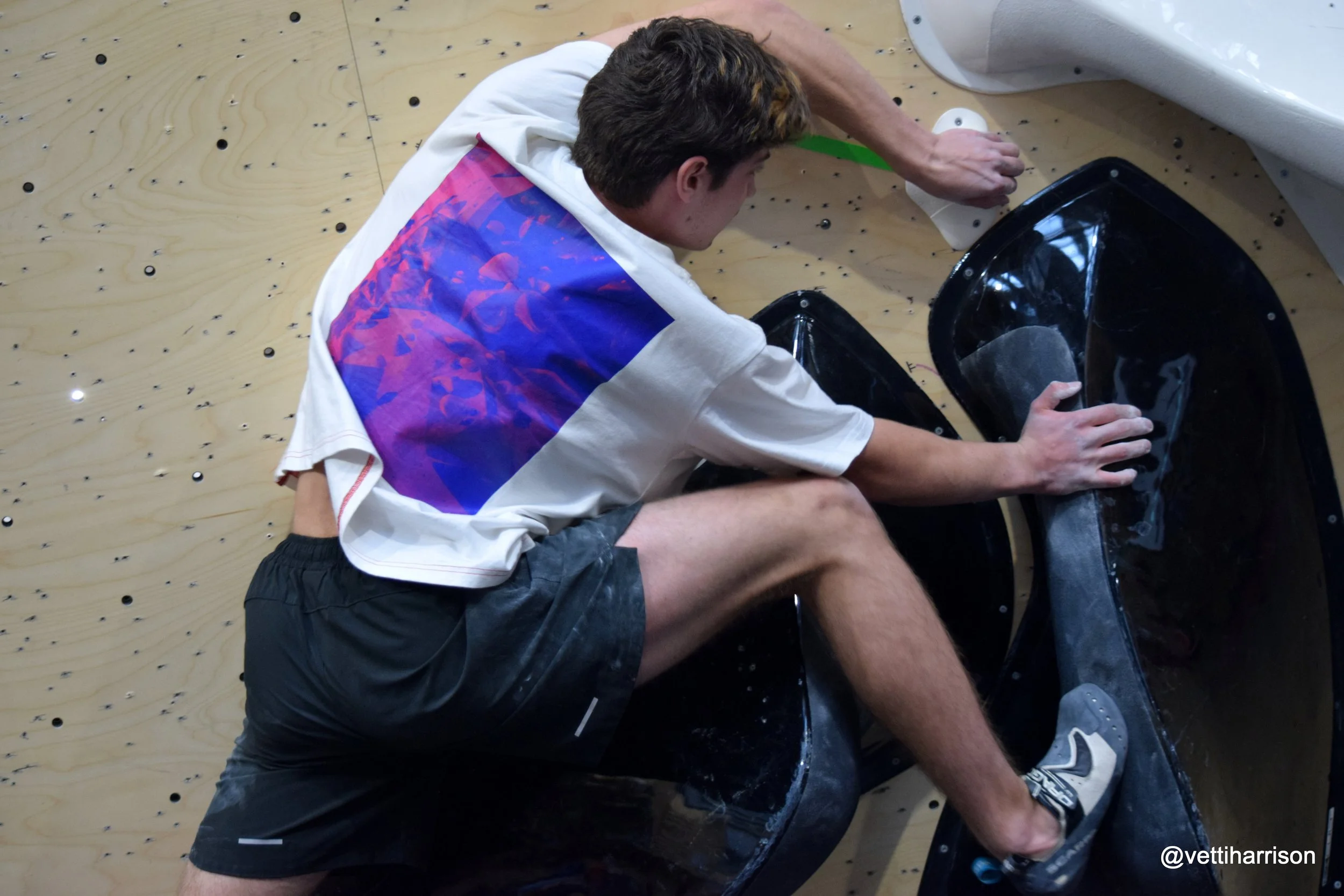 Person with brown hair climbing a rock wall, wearing a white shirt with a blue and purple graphic on the back, and dark shorts.