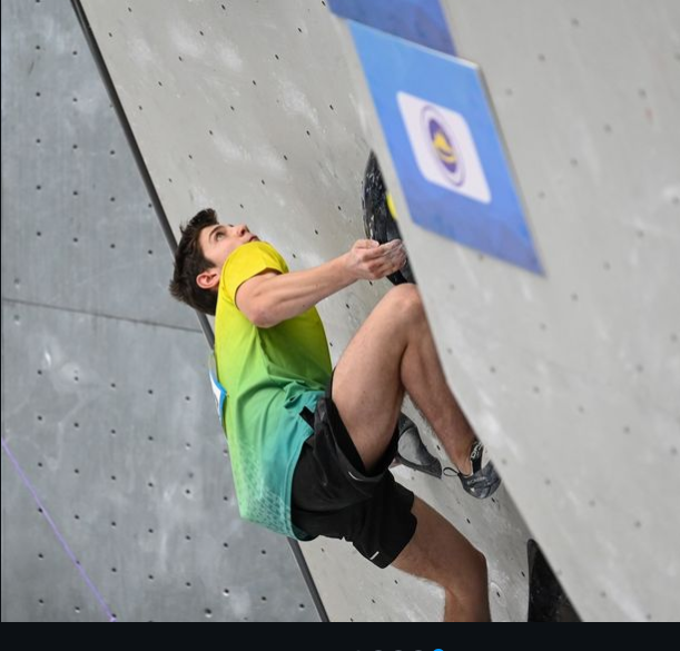 Young male climber wearing a yellow and green shirt, black shorts, and climbing shoes, scaling an indoor rock climbing wall with gray holds.