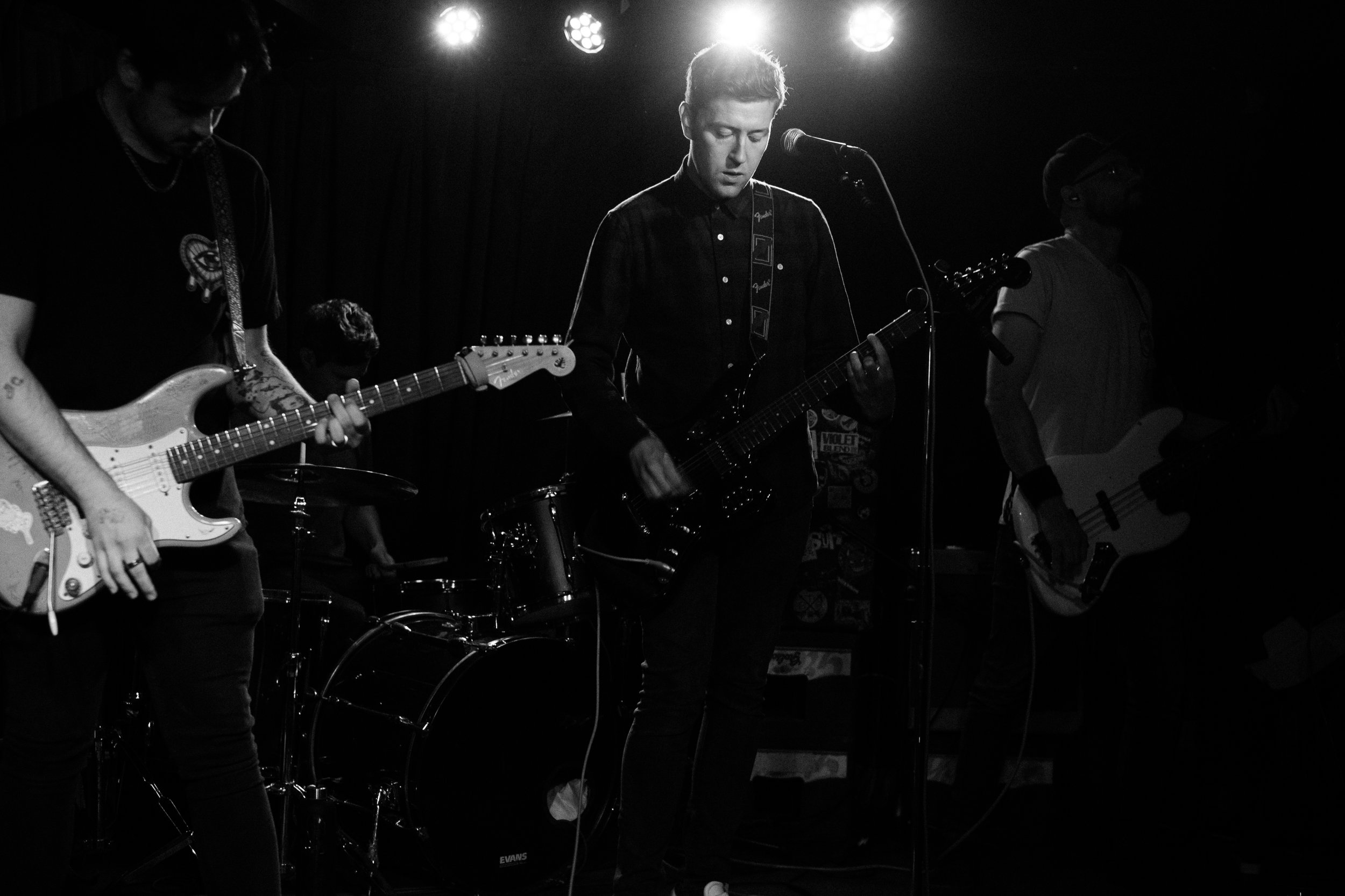 Black and white photo of a band playing live music on stage, featuring four musicians with guitars and drums, illuminated by stage lights.