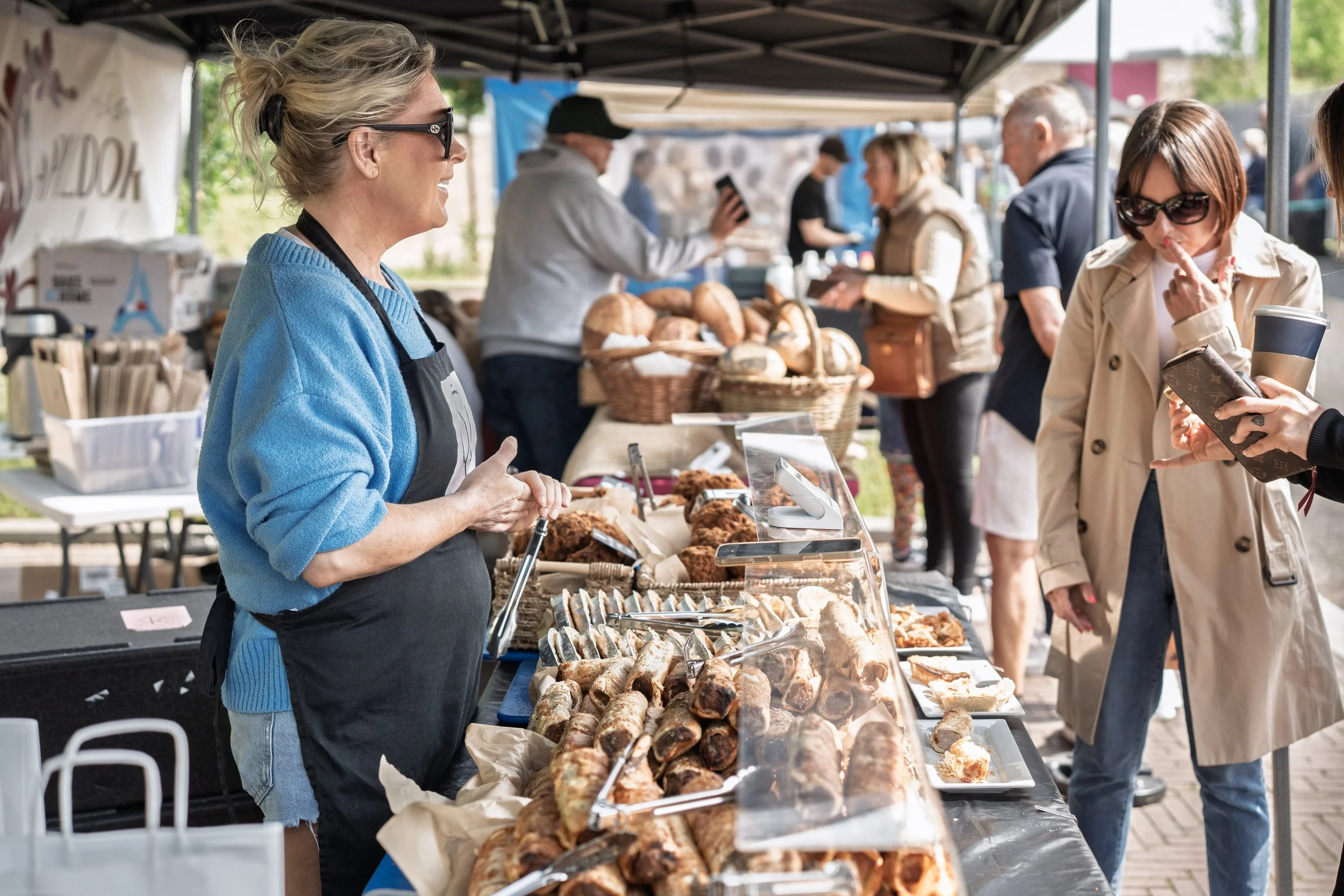 A woman wearing sunglasses, a blue sweater, and an apron stands behind a food stall selling baked goods at an outdoor market. Several customers are browsing and purchasing items, with baked goods displayed on the table, including rolls and pastries.