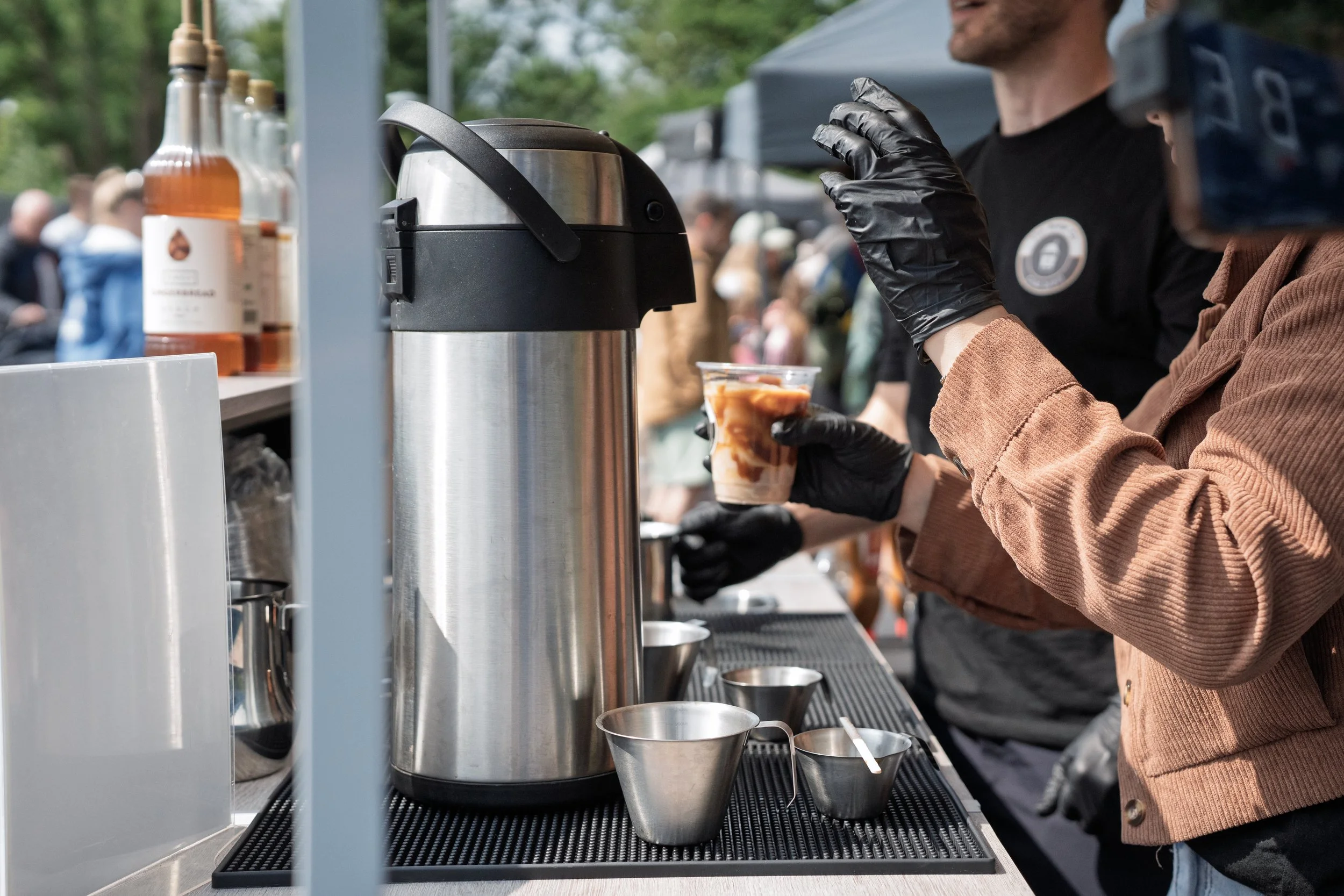 A person wearing a black glove and a brown jacket is serving a drink from a thermal coffee urn at an outdoor event. There are bottles of flavored syrup and other people in the background.