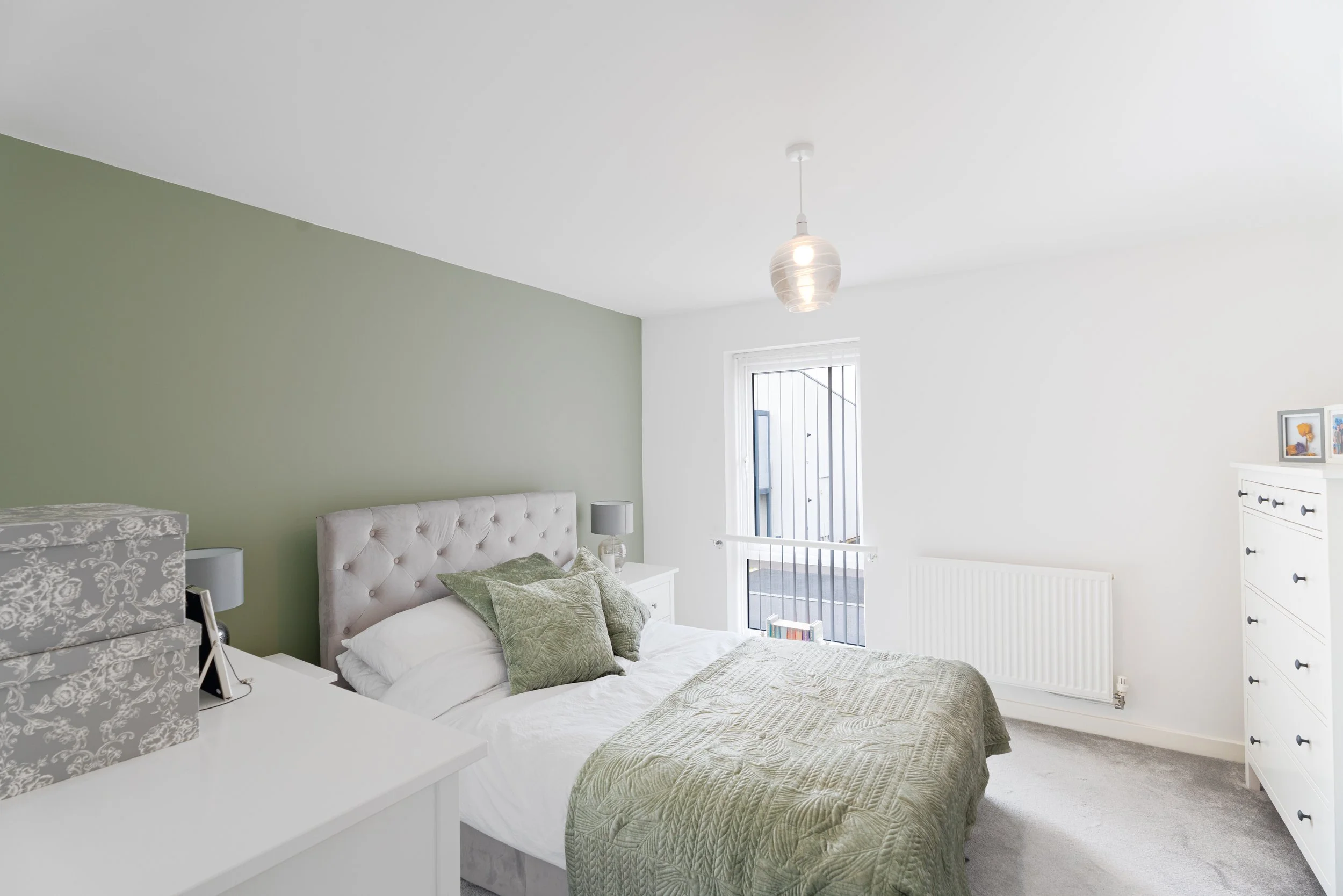 A bedroom with a white bed, green quilt, and pillows, a white dresser with framed pictures, a white nightstand with a lamp, and a window with bars, featuring a light fixture hanging from the ceiling.