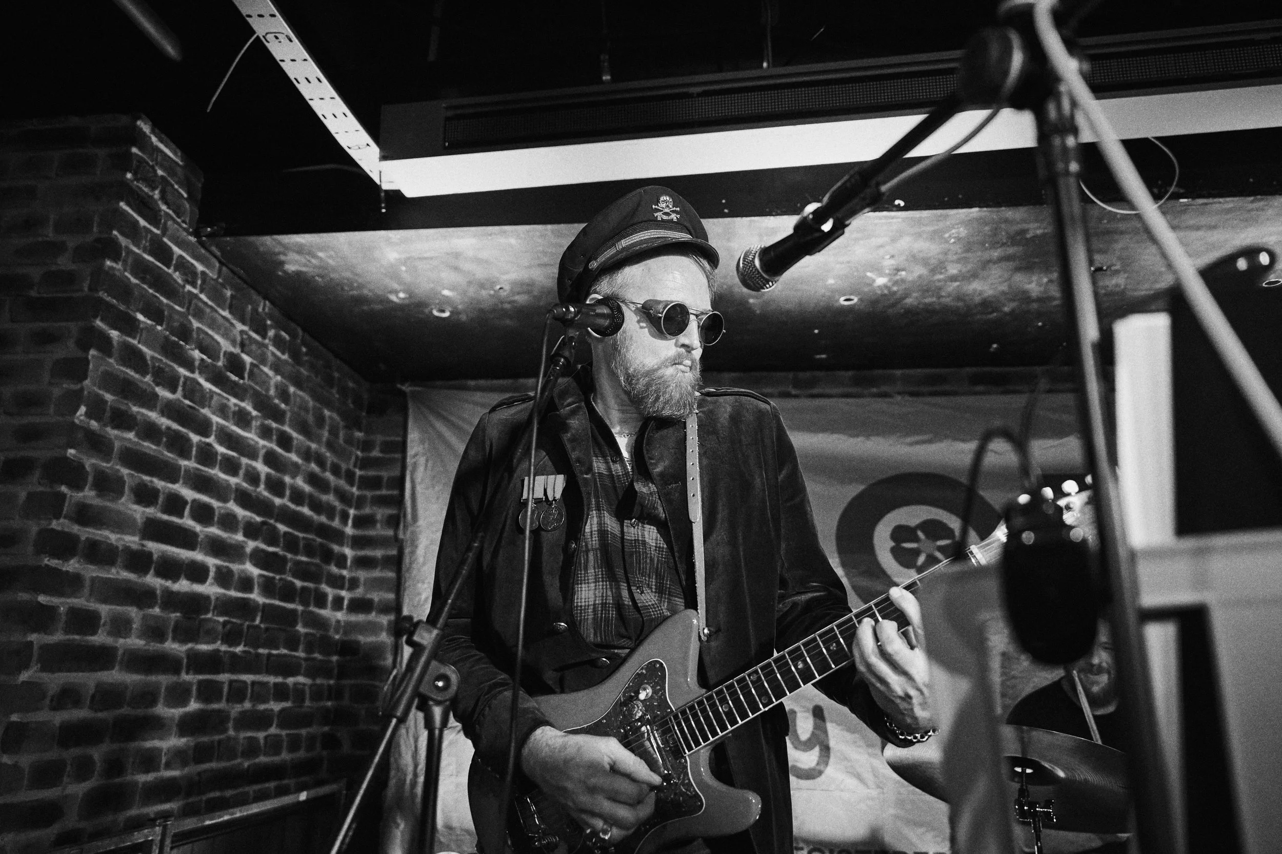 A man wearing a hat and sunglasses playing an electric guitar and singing into a microphone in a music venue with brick walls.