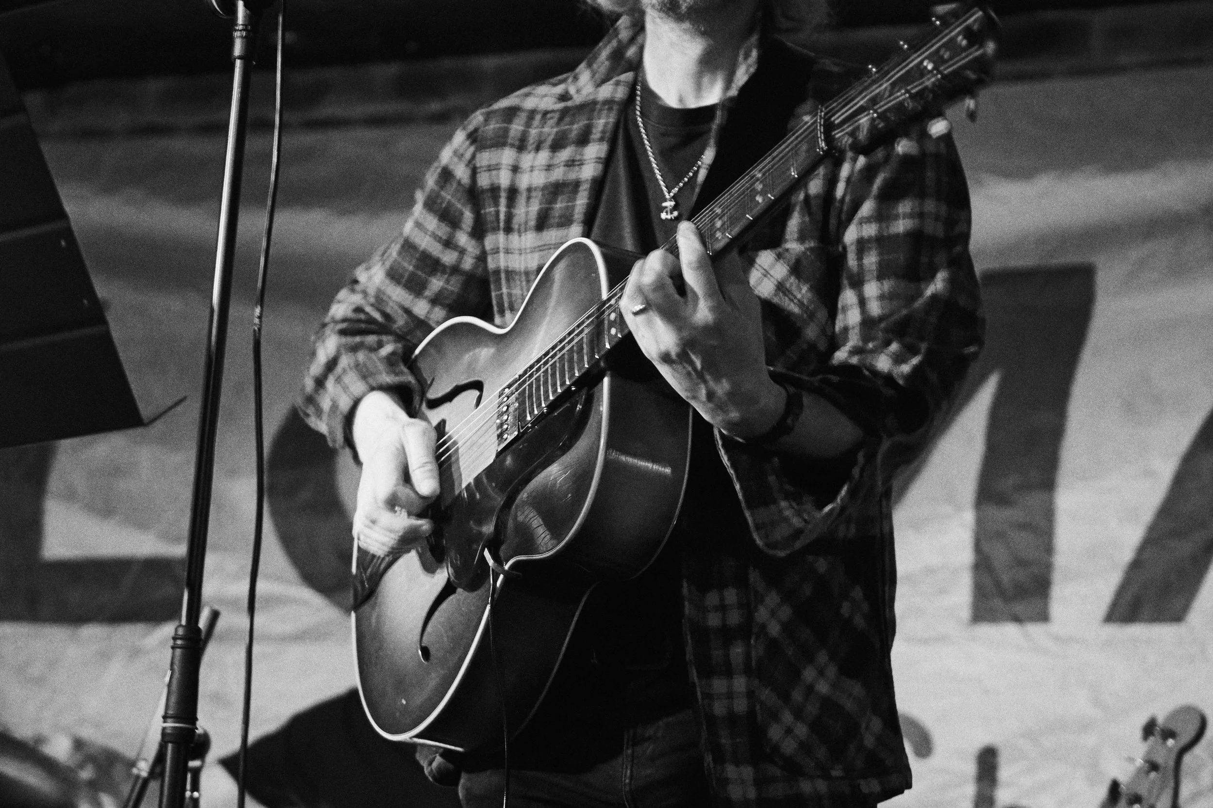 A person playing an acoustic guitar in a black and white photo, wearing a checkered shirt and a necklace, with a microphone stand nearby.