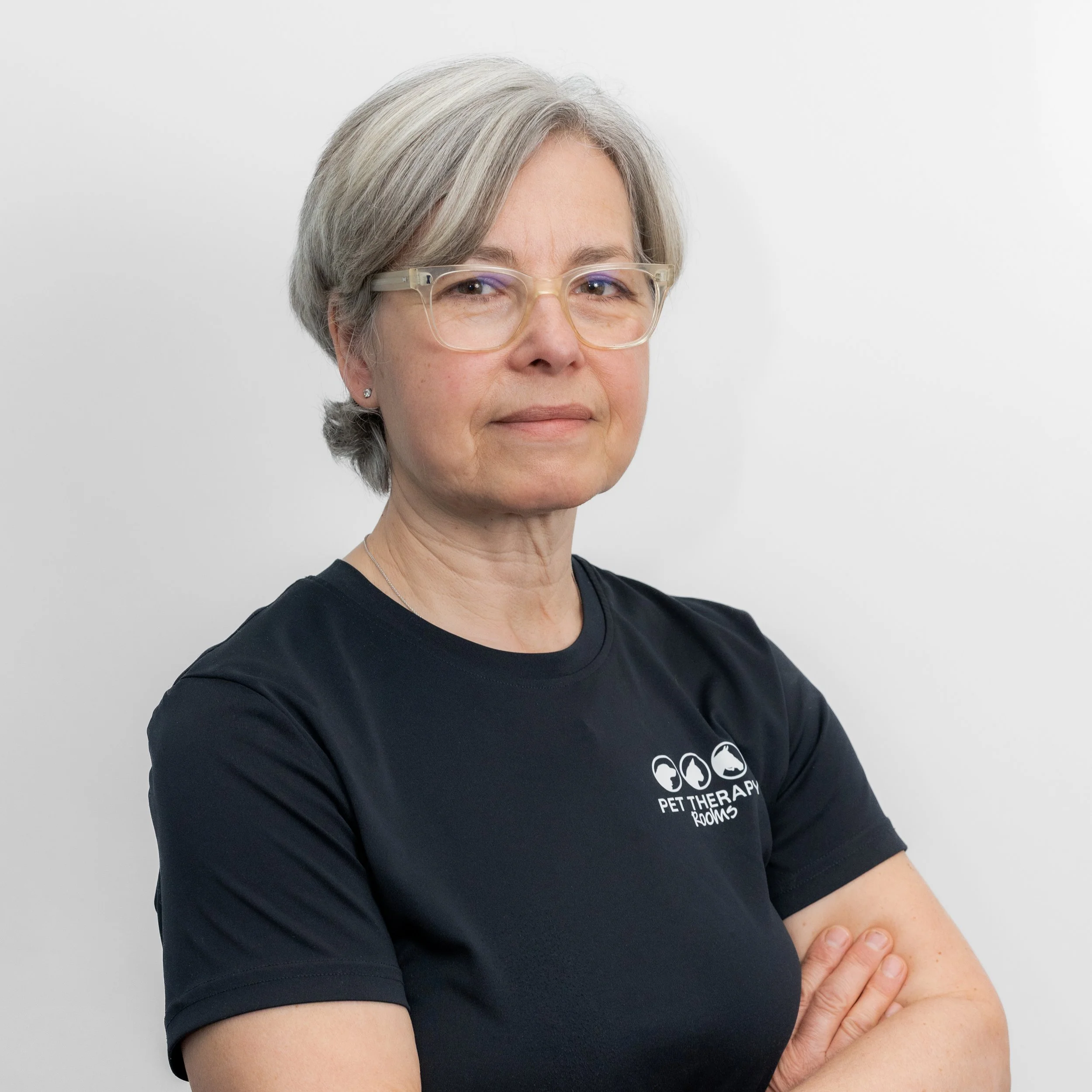 A woman with short gray hair and glasses, wearing a black t-shirt with a logo which reads pet therapy rooms. She is standing against a plain white background, folding her arms.
