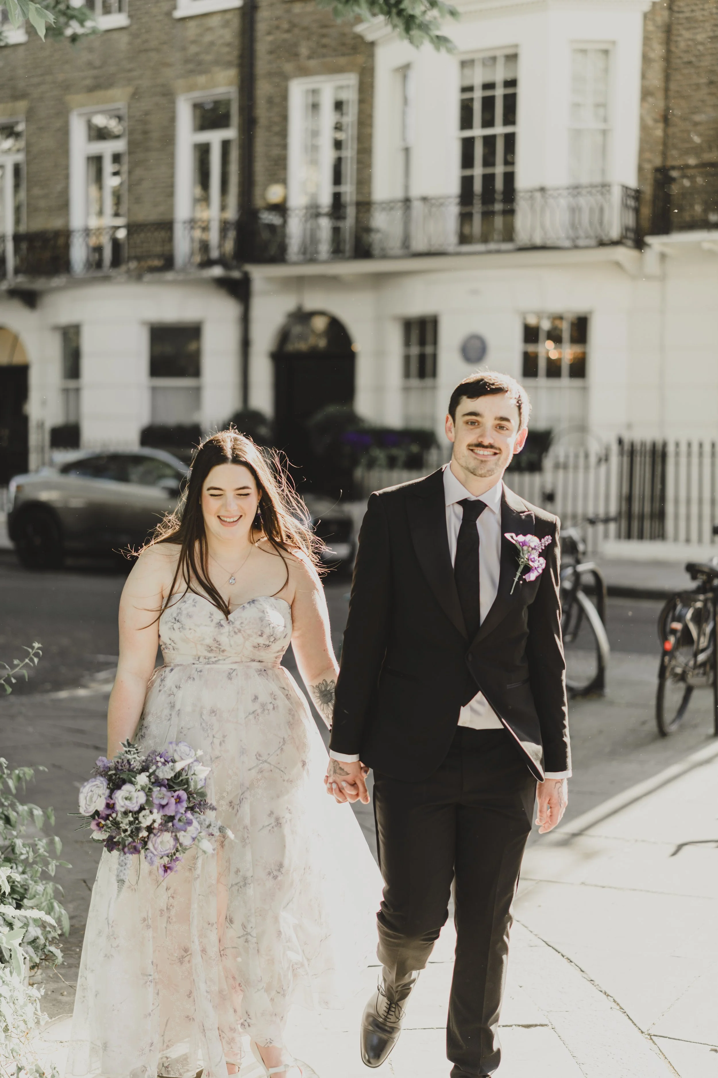 A smiling couple holding hands, walking outside on a city street. The woman wears floral dress and holds a purple flower bouquet. The man is dressed in a black tuxedo, white shirt, and black tie, with a purple boutonniere. The background shows buildings and parked cars.