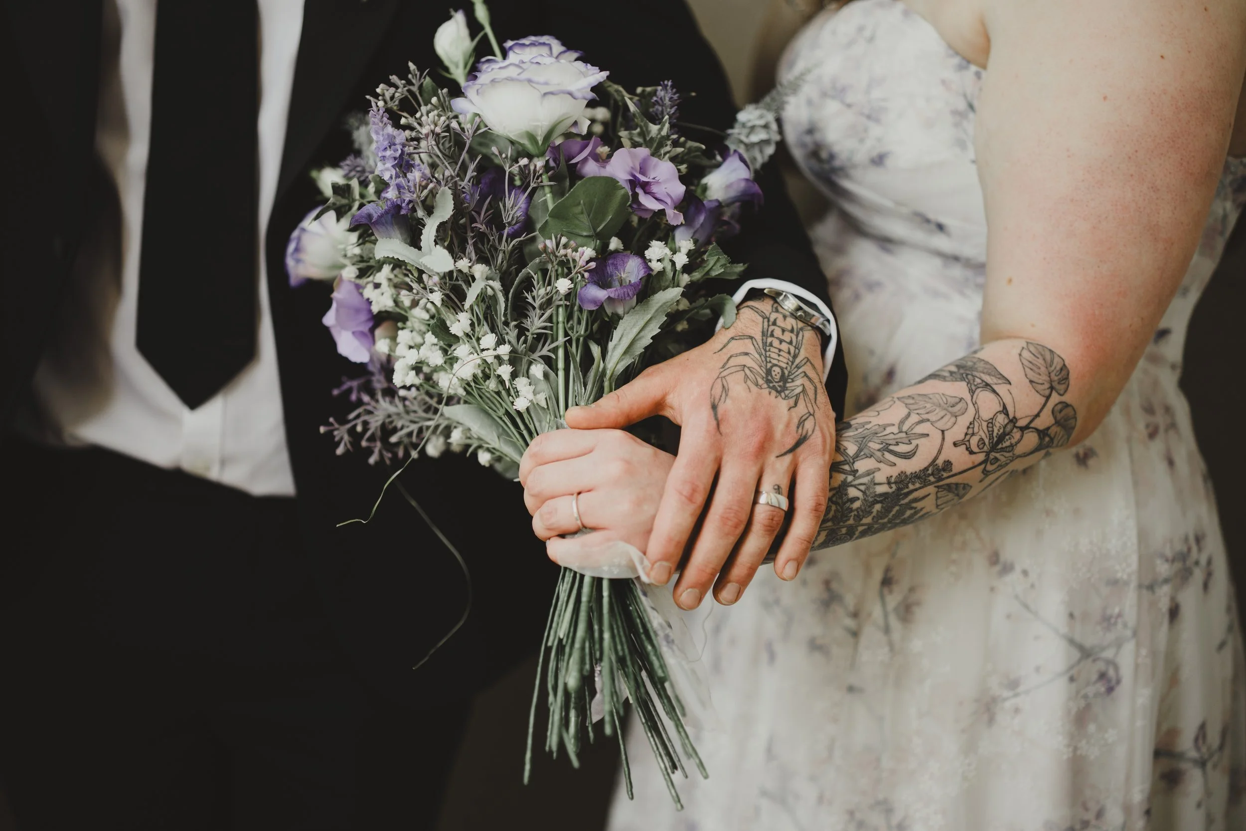A bride and groom with tattoos on their arm holding a bouquet of purple, white, and lavender flowers at a wedding ceremony.