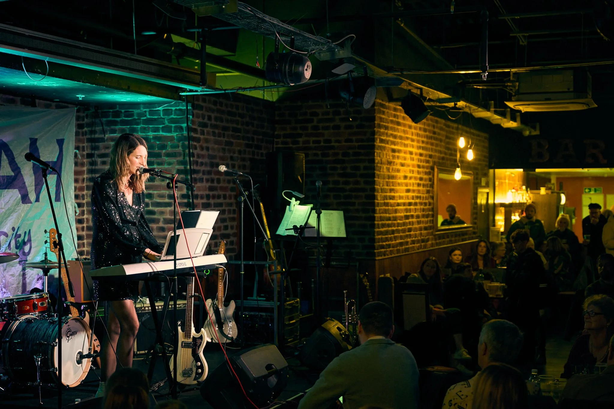 A woman playing keyboard on stage at a live music event held in a venue with brick walls and warm lighting, with an audience seated and watching her performance.