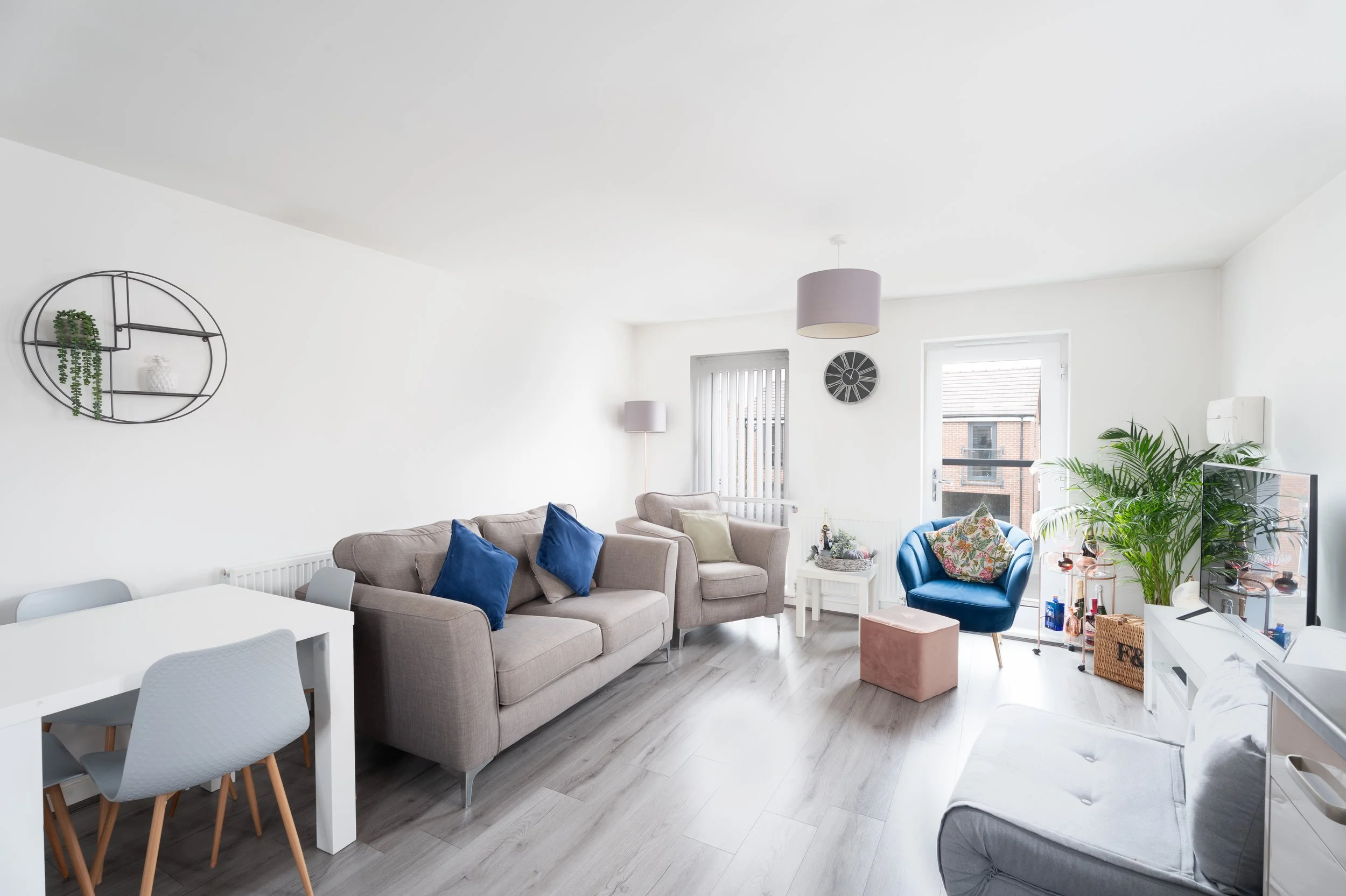 Living room with beige sofa, blue armchair, white walls, and window letting in natural light. Decor includes wall clock, shelves, plants, and various furniture and decorative items.