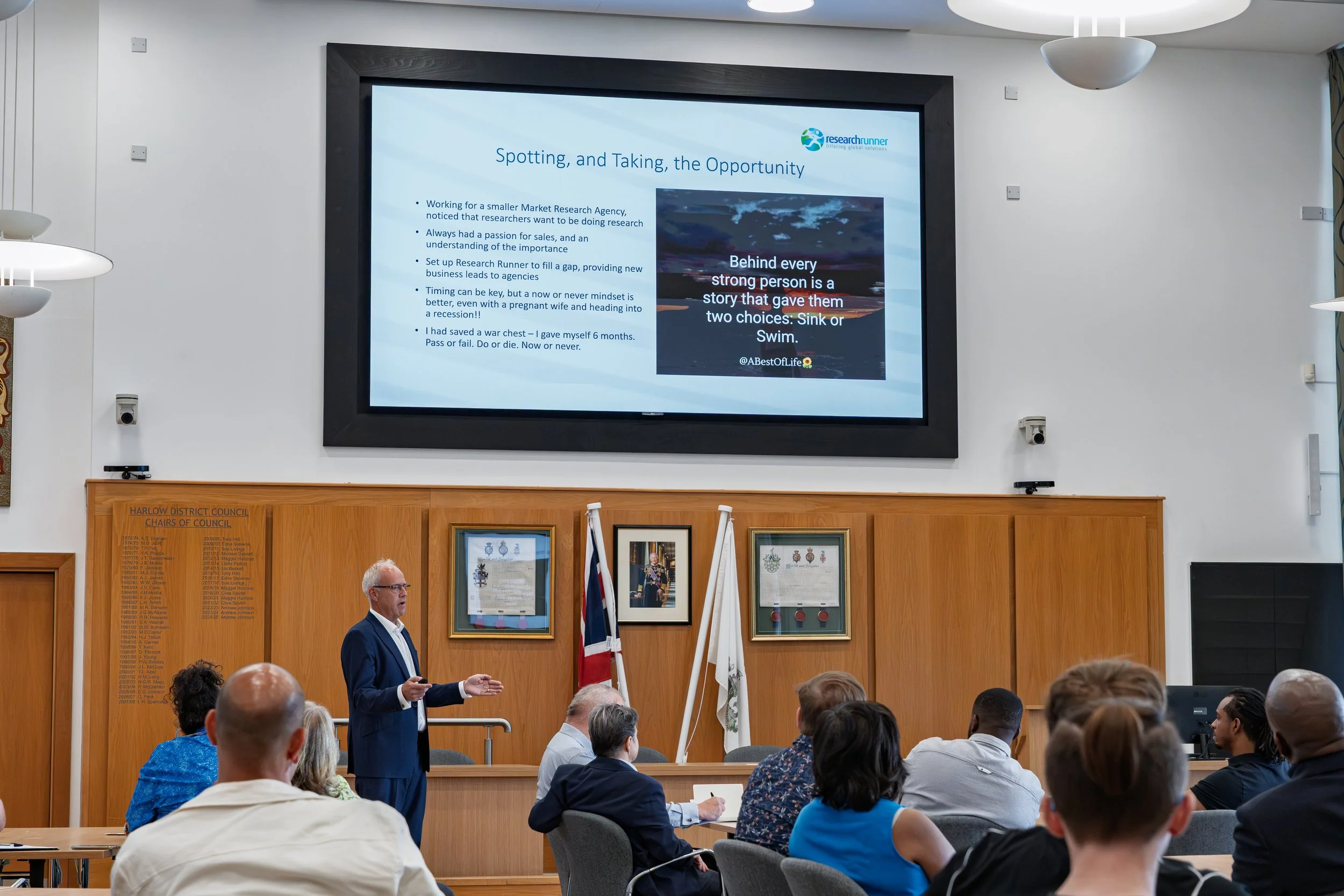 A man in a suit giving a presentation to an audience in a conference room with a large screen displaying a slide titled 'Spotting, and Taking, the Opportunity'. The audience members are seated facing the presenter. The room has wooden paneling and fr