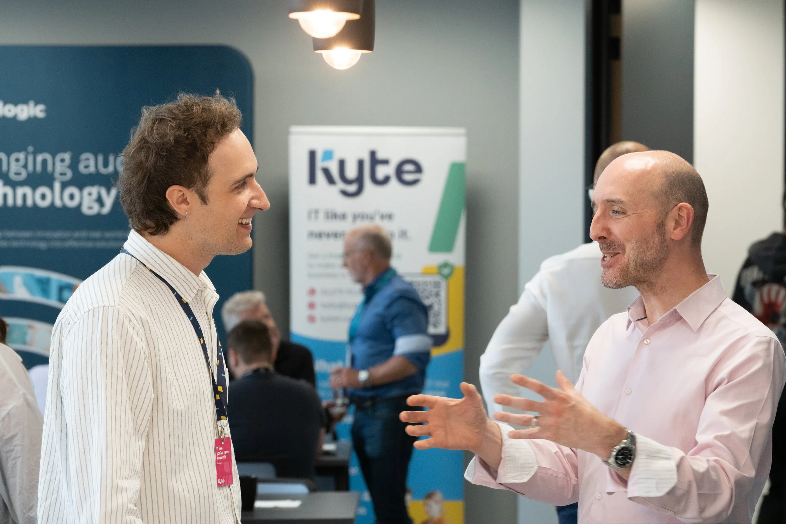Two men are engaged in conversation at a professional conference or event. The man on the left is smiling and wearing a striped collared shirt with an event badge, while the man on the right is speaking and gesturing with his hands, dressed in a ligh