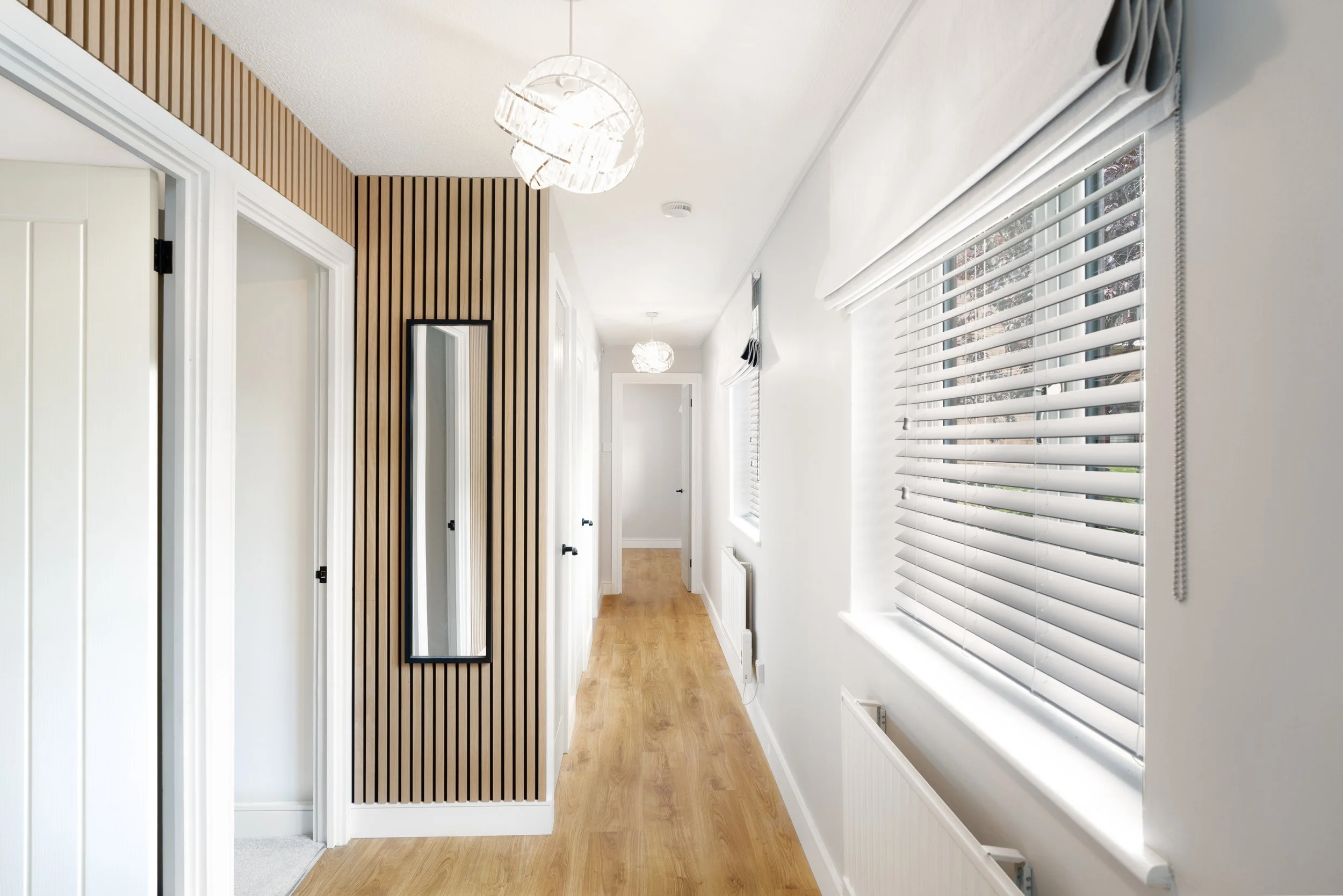 A bright hallway with white walls, wooden flooring, and large windows with white blinds on the right side. There are three pendant lights with white shades hanging from the ceiling. A full-length mirror is mounted on a wall with vertical wooden slats