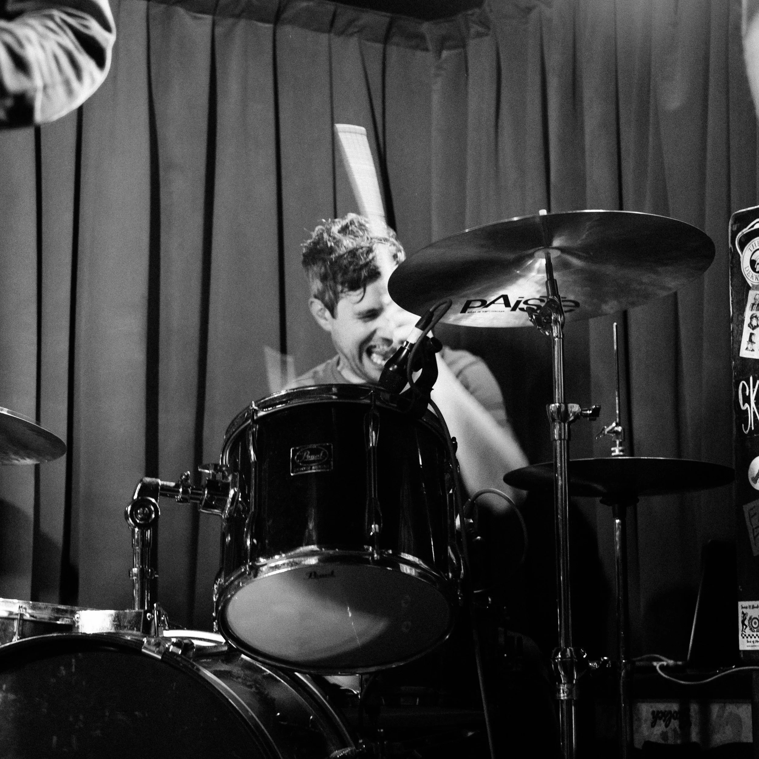A black and white photo of a young man with curly hair playing a drum kit, smiling, with a wooden wall in the background.