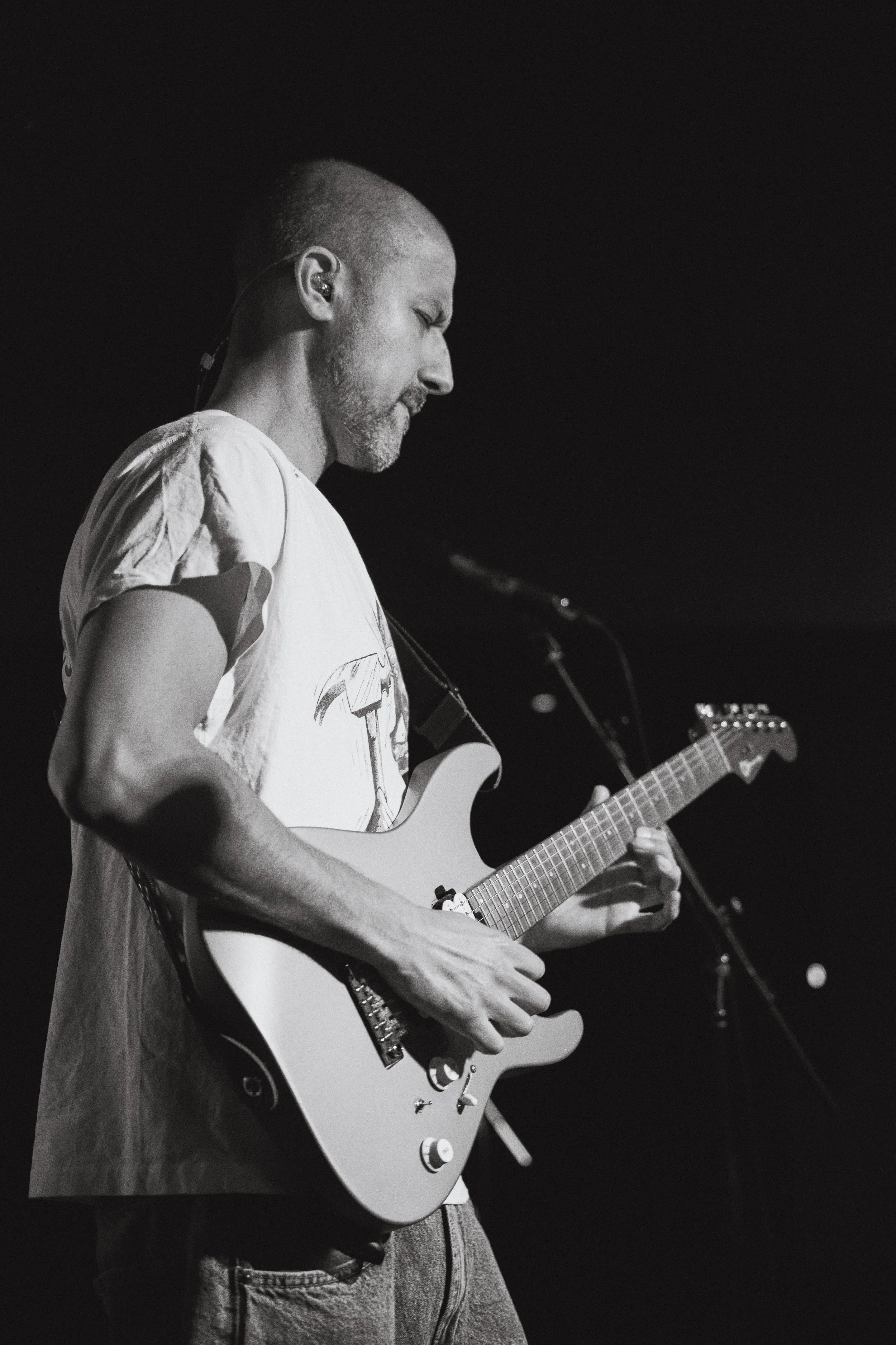 Black and white image of a man with a shaved head playing an electric guitar, with eyes closed, possibly performing on stage.