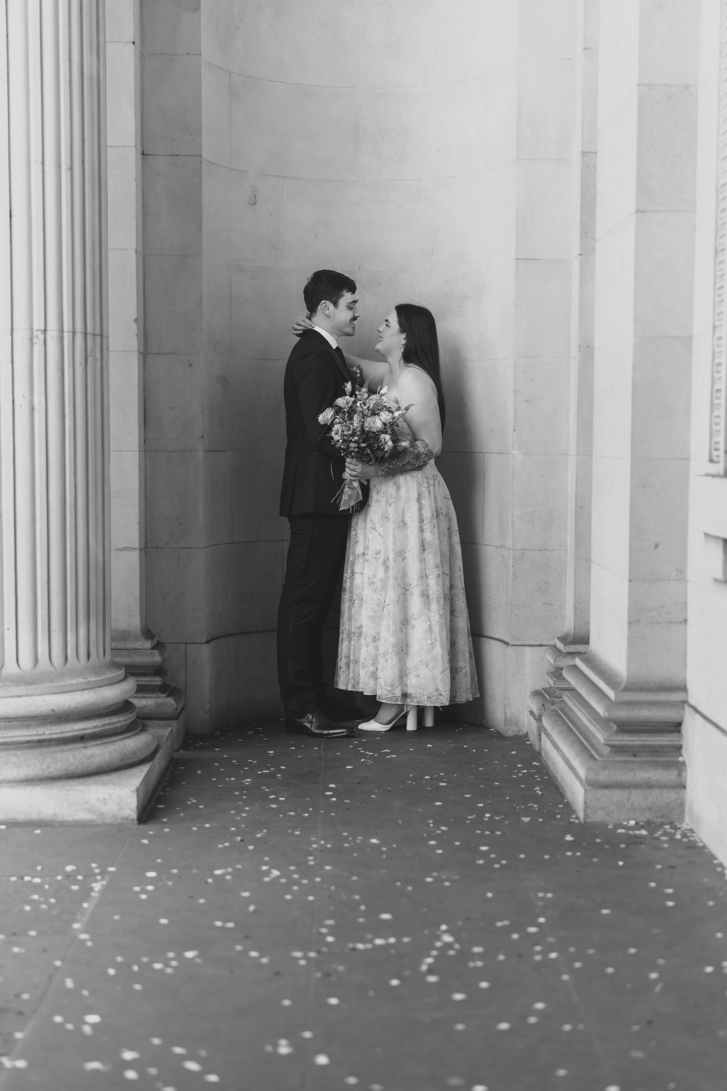 A black-and-white photo of a couple in wedding attire standing close to each other, holding a bouquet, and gazing into each other's eyes between large classical columns with a stone wall background.