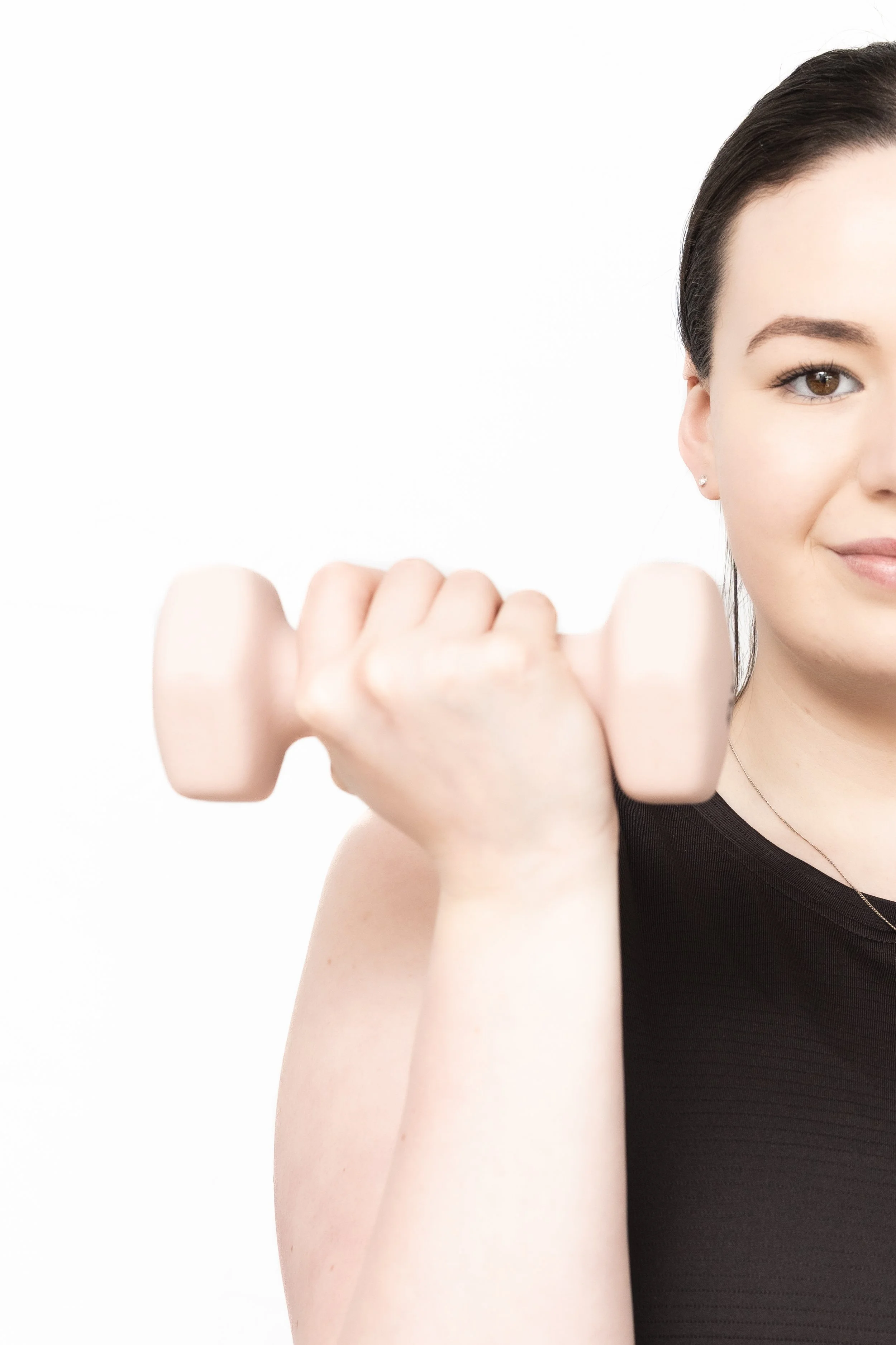 A young woman with dark hair styled back, wearing a black tank top, holding a small light pink dumbbell in front of her with one hand, smiling slightly, against a plain white background.