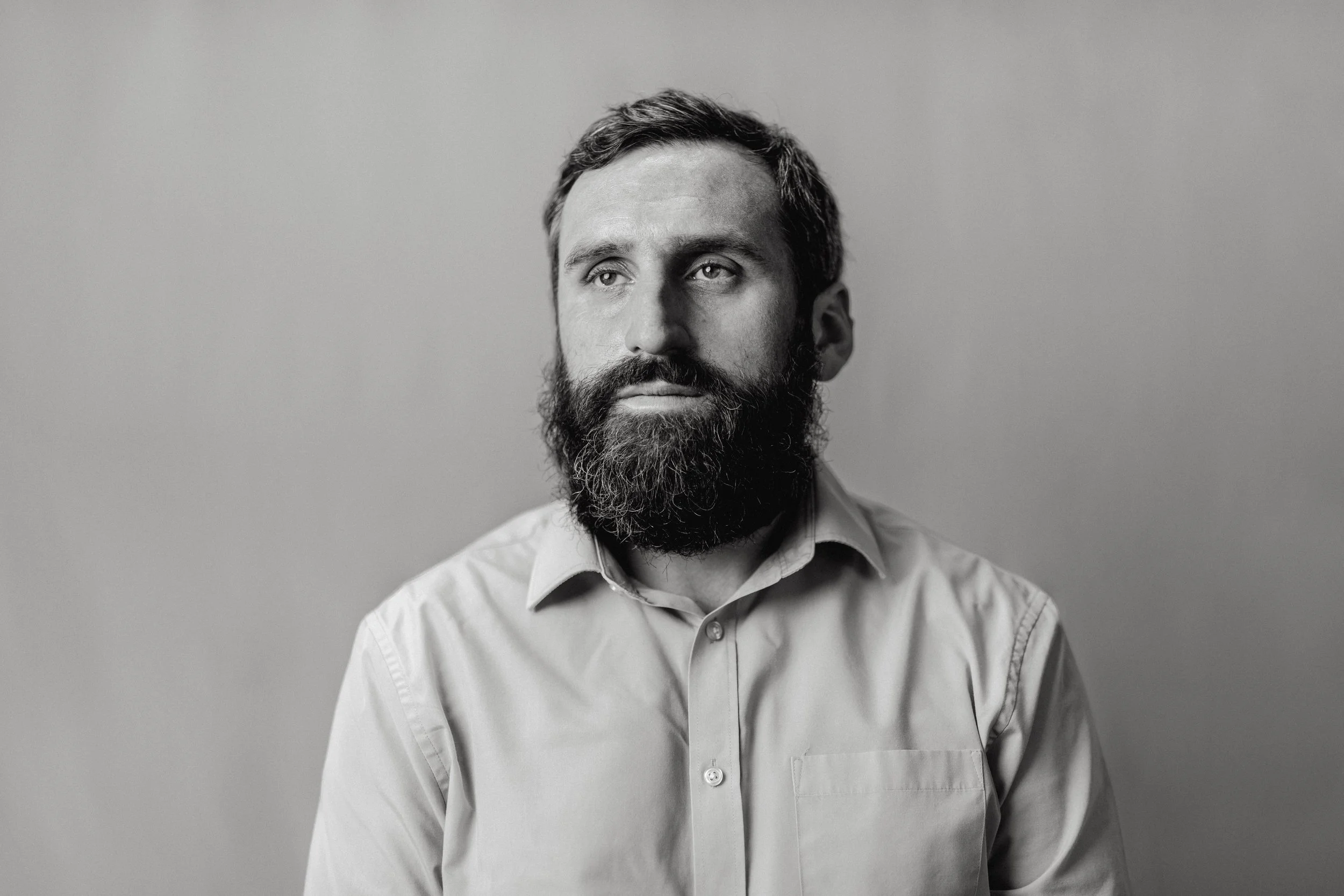 Black and white portrait of a man with a beard, wearing a collared shirt, looking slightly away from the camera against a plain background.