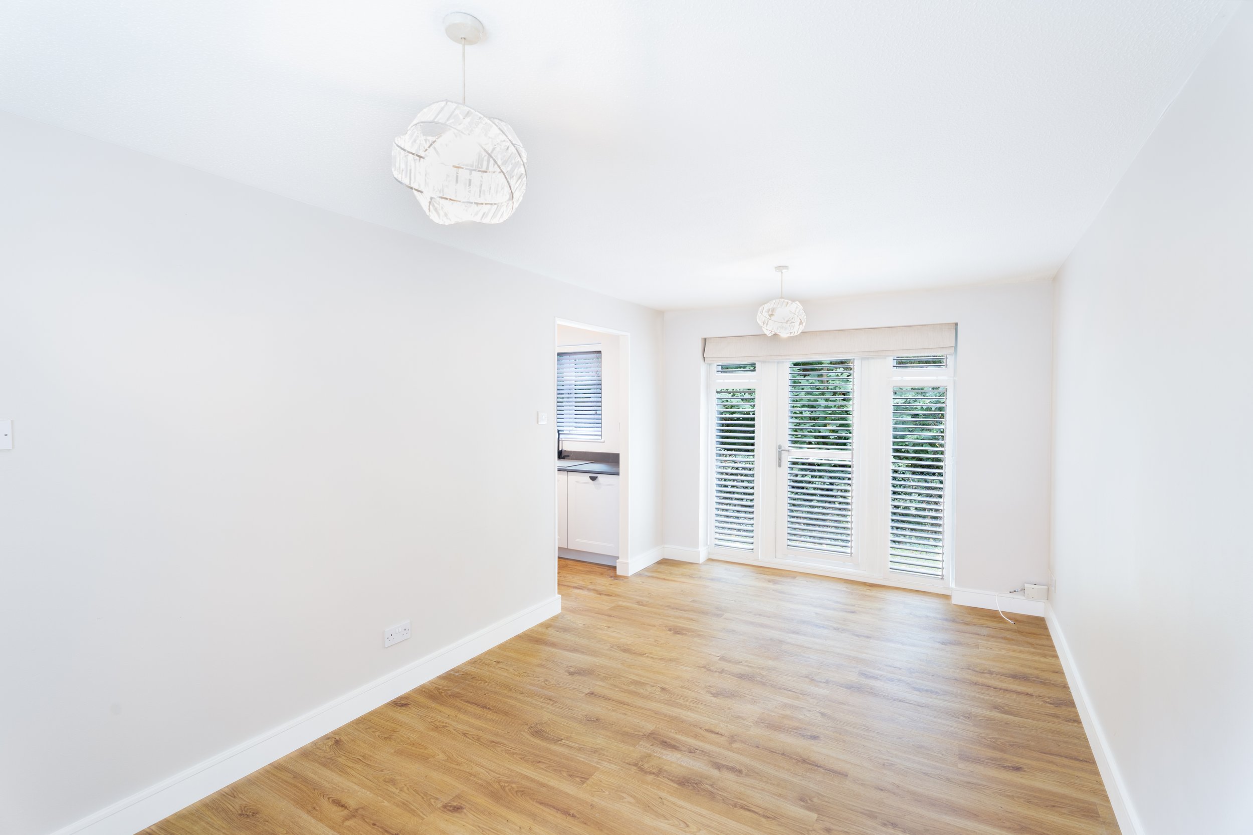 Empty living room with white walls, hardwood floor, large windows with blinds, and ceiling light fixtures.