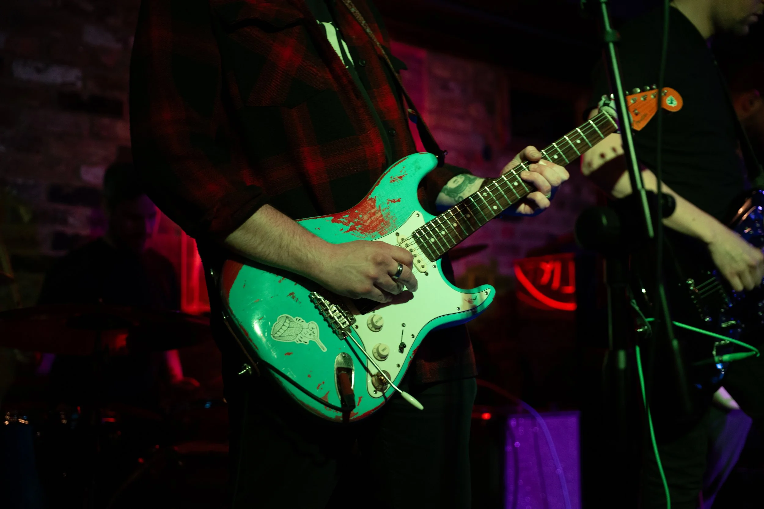 A person playing an electric guitar on stage with a brick wall background, wearing a black and red checkered shirt, with colored stage lighting.