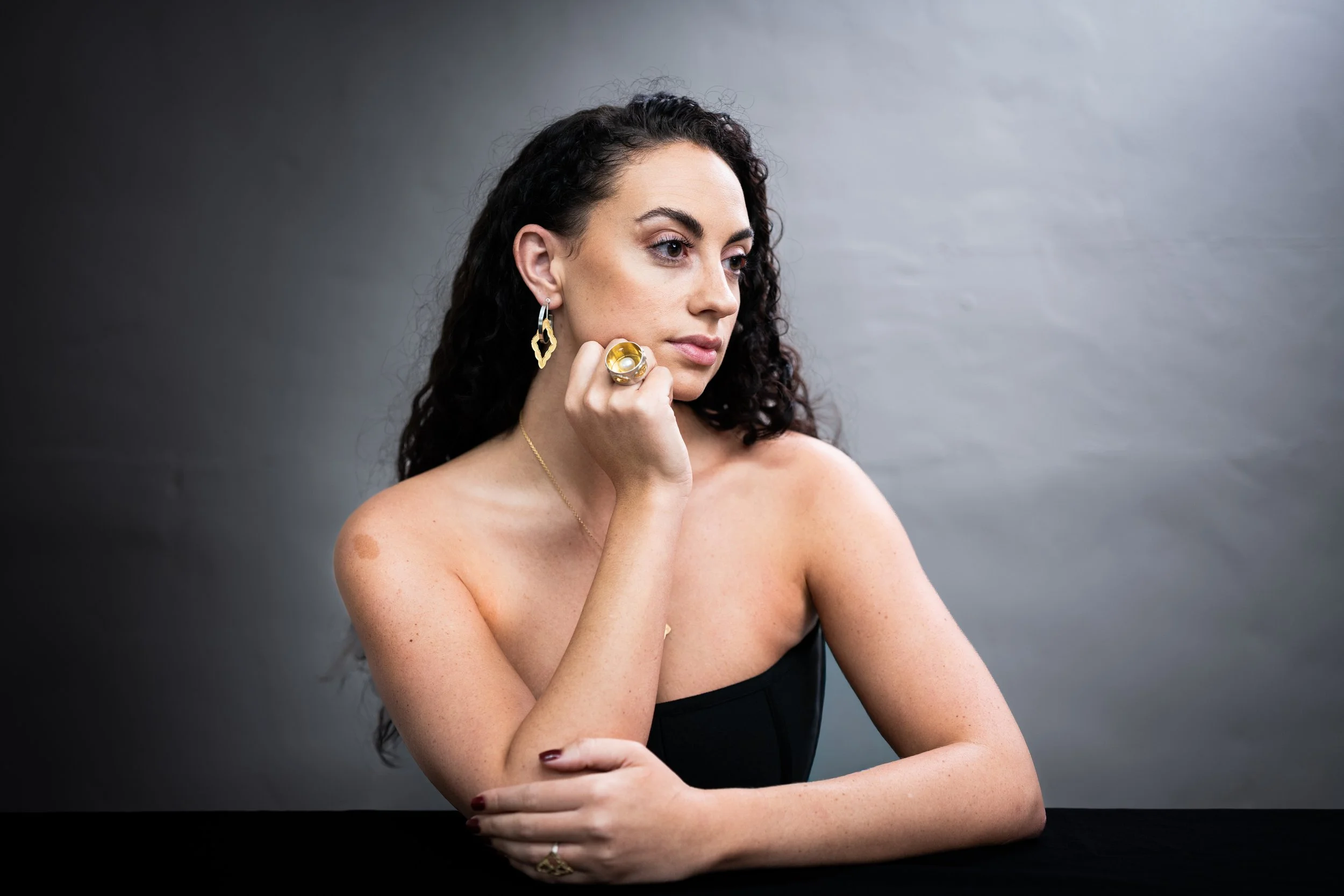 A woman with curly dark hair and earrings looking to the right, resting her chin on her hand, wearing a black strapless top, against a gray background.