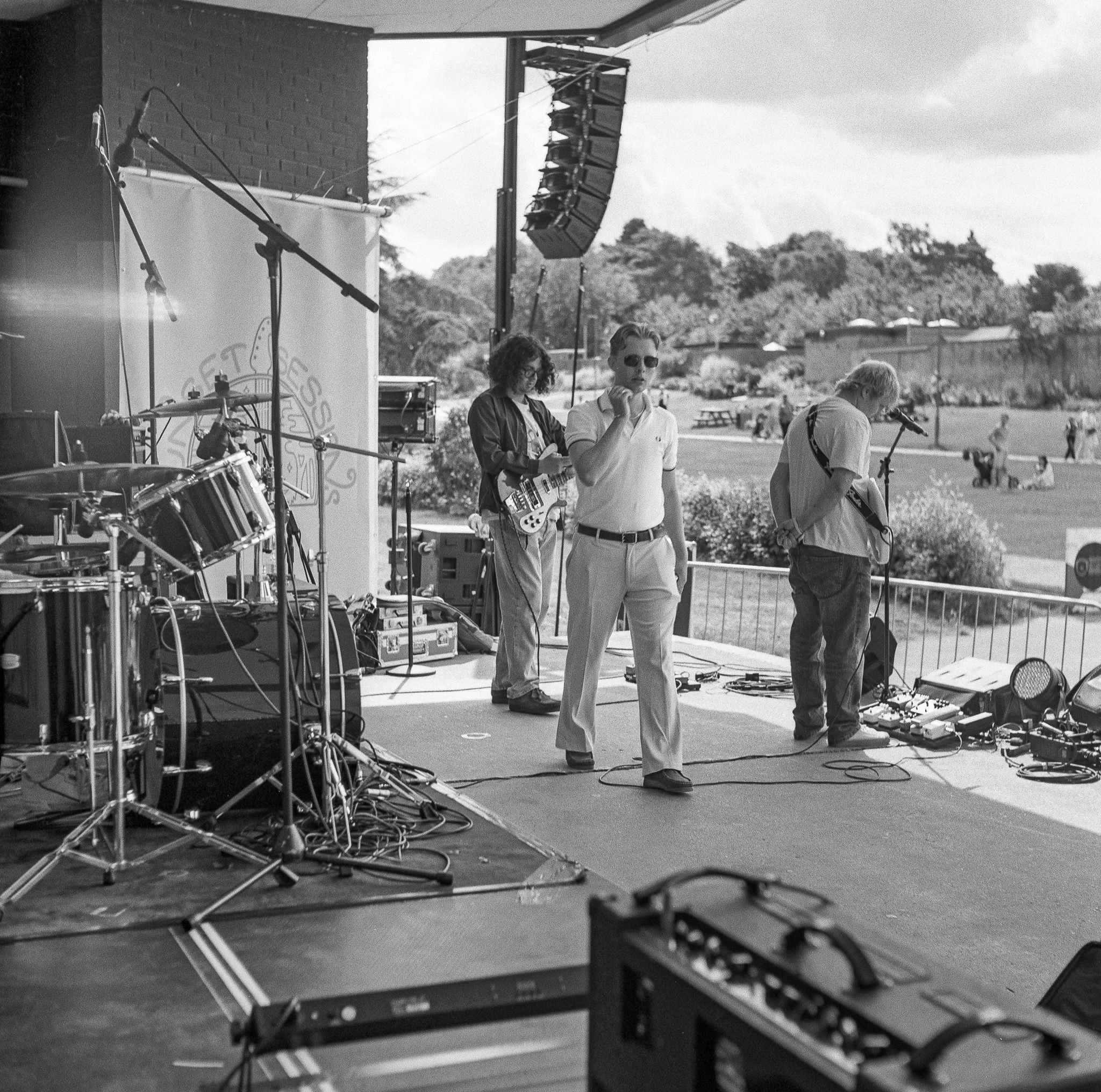 Black and white photo showing an outdoor music performance with musicians, microphones, and sound equipment on stage, with a scenic background of trees and houses.