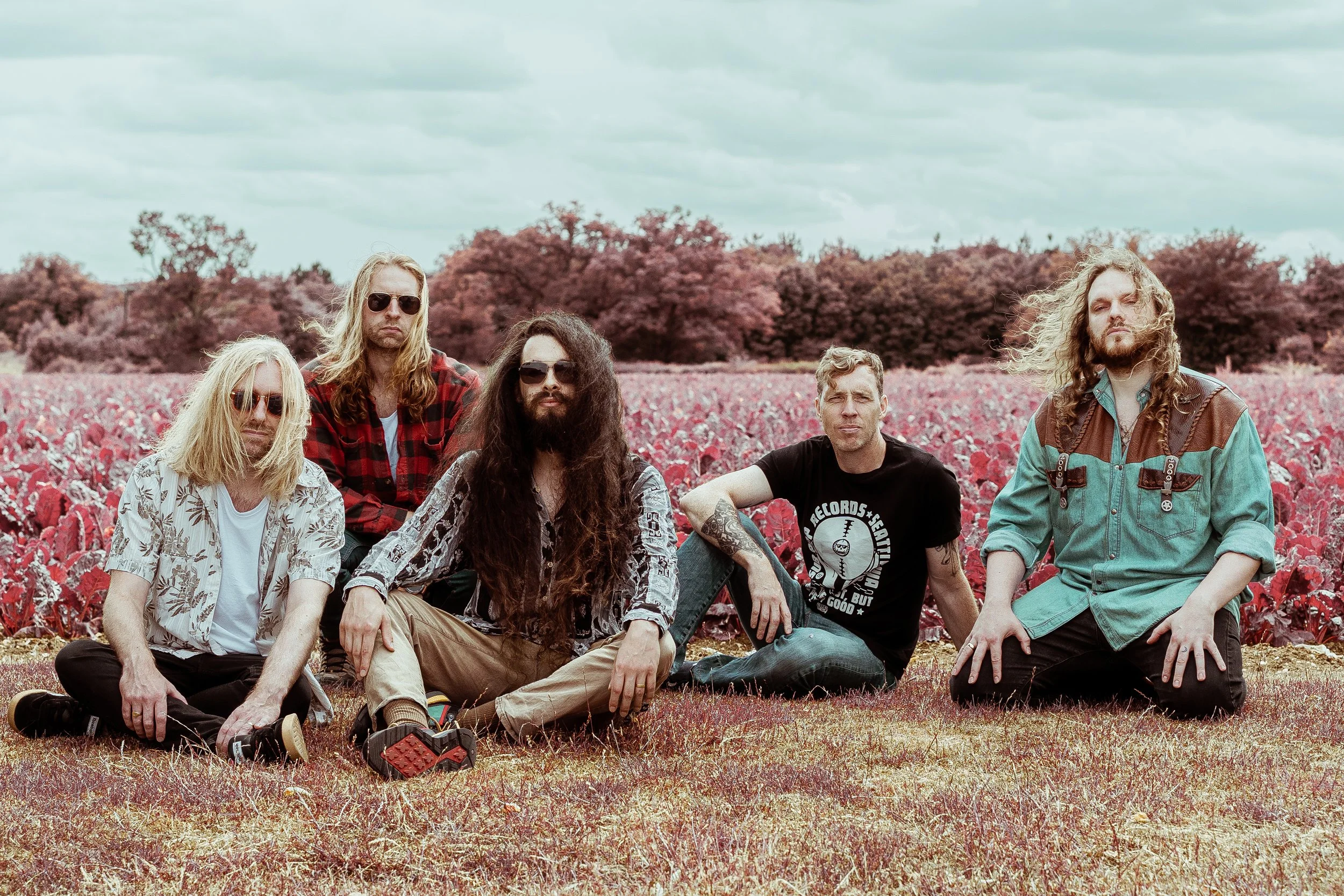 A group of six young men with long hair, some wearing sunglasses, sitting and kneeling on a field with an aerochrome effect. 