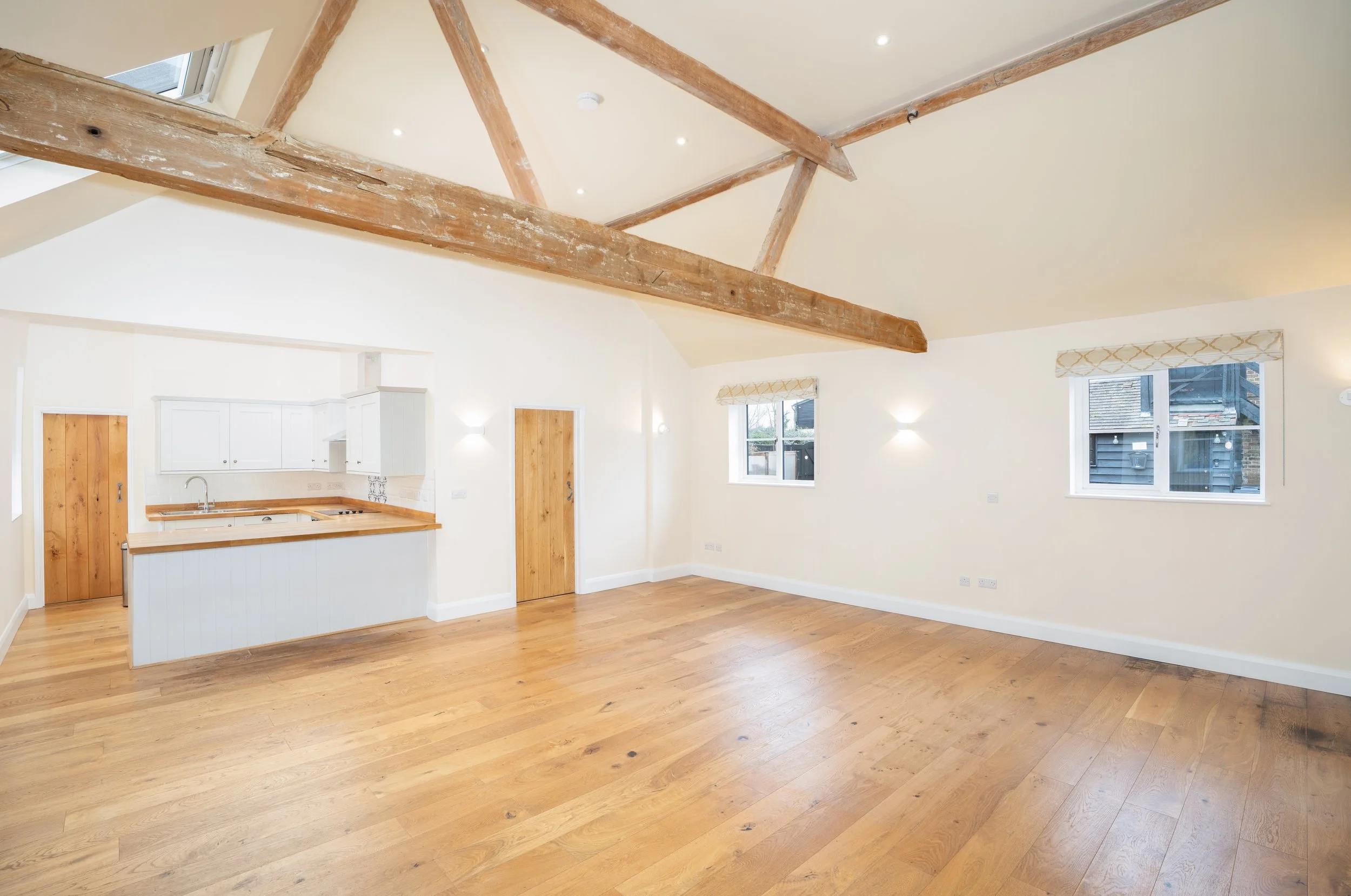 Spacious, open-concept living area with exposed wooden beams, large windows, and hardwood floors, connected to a kitchen with white cabinets and wooden accents.