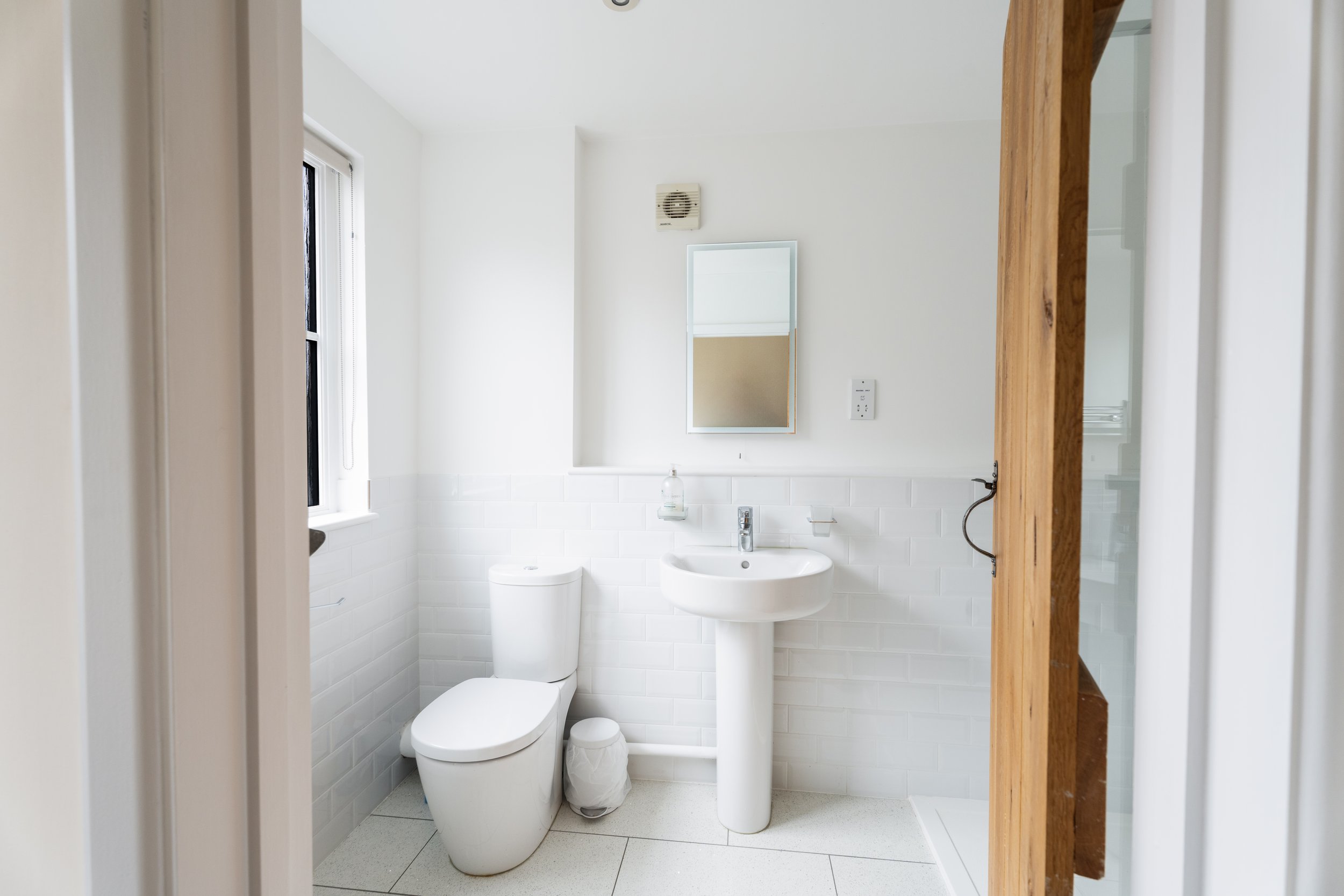 A small, bright bathroom with a white toilet, white sink, and a mirror. The walls are painted white with a half-tile wainscot. There's a small window on the left and a ventilation fan on the wall. A wooden door frame on the right partially shows the 