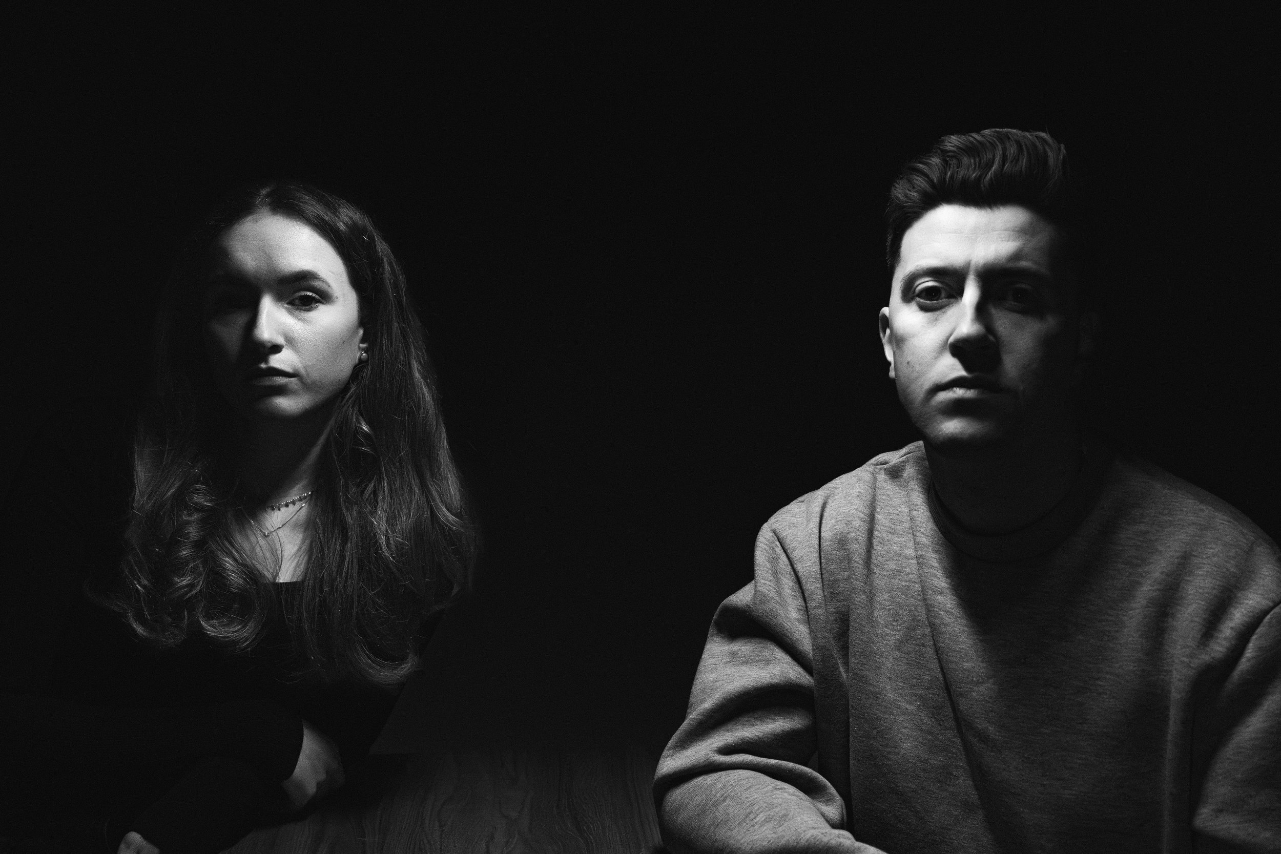 Black and white portrait of a woman with long, wavy hair on the left and a man with short, styled hair on the right, both looking directly at the camera with serious expressions.