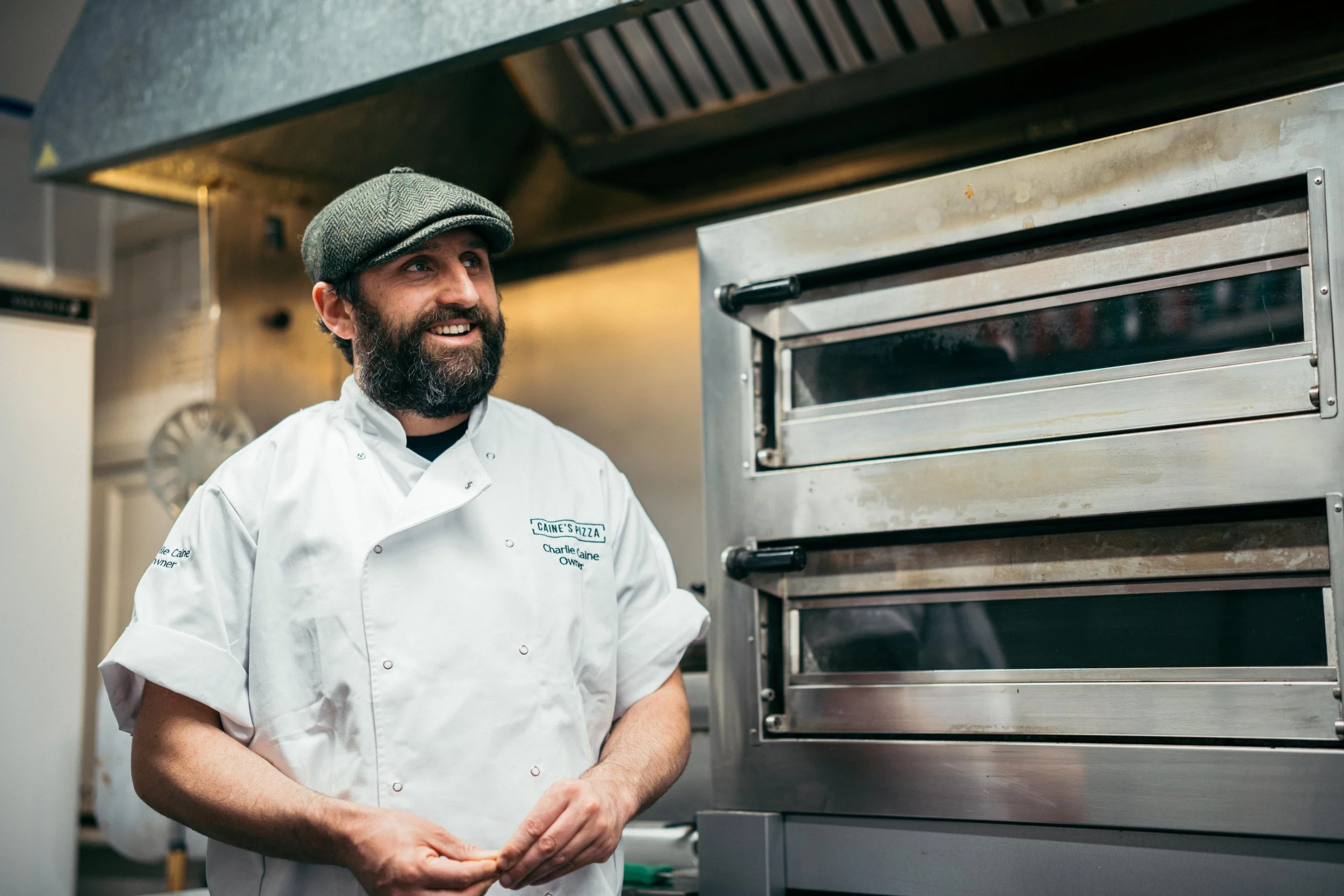 A man with a beard and a flat cap, wearing a white chef's coat, standing in a commercial kitchen near a metal oven, smiling and looking away from the camera.