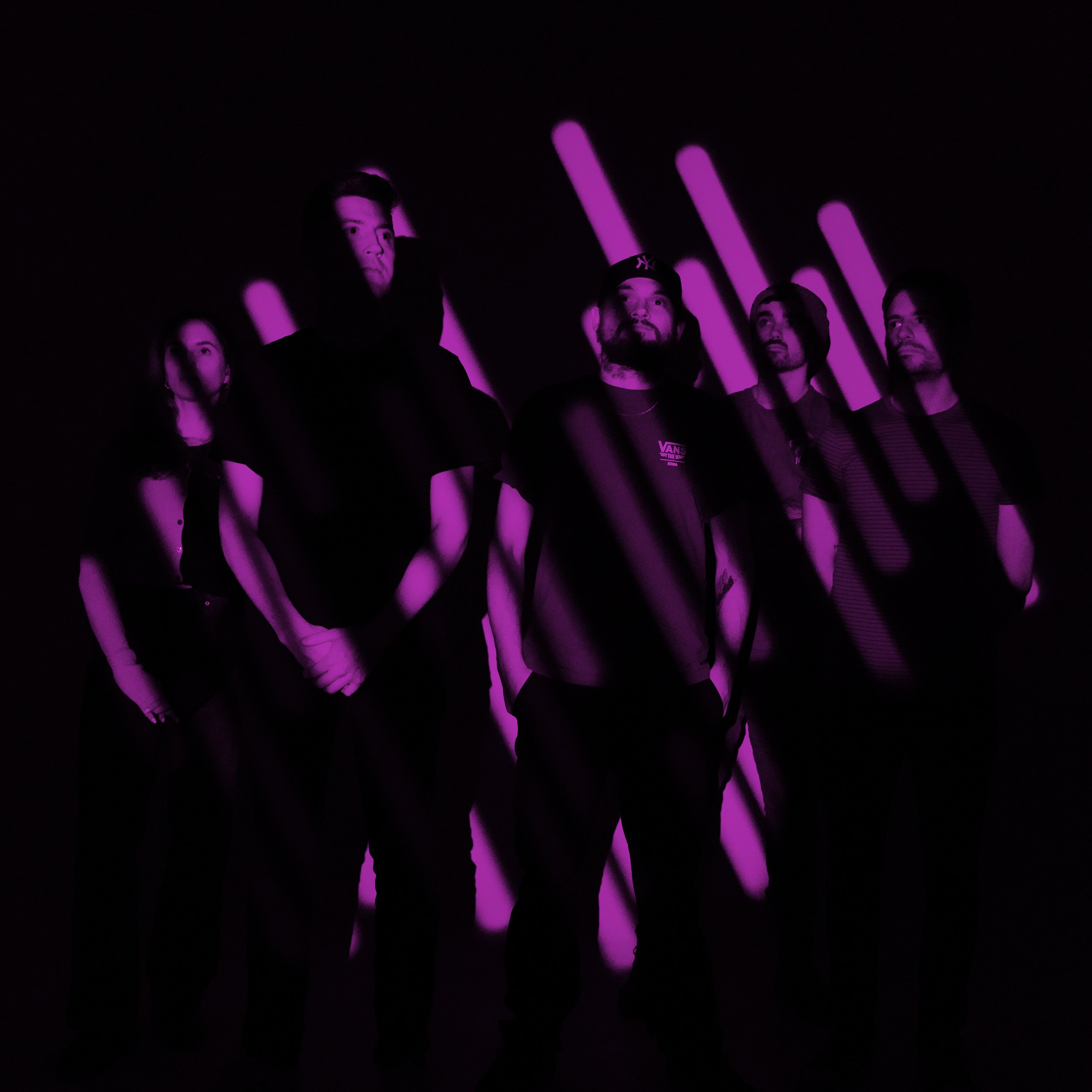 Group of people standing in front of pink and purple light bars, some wearing hooded sweatshirts and a baseball cap, with a dark background.