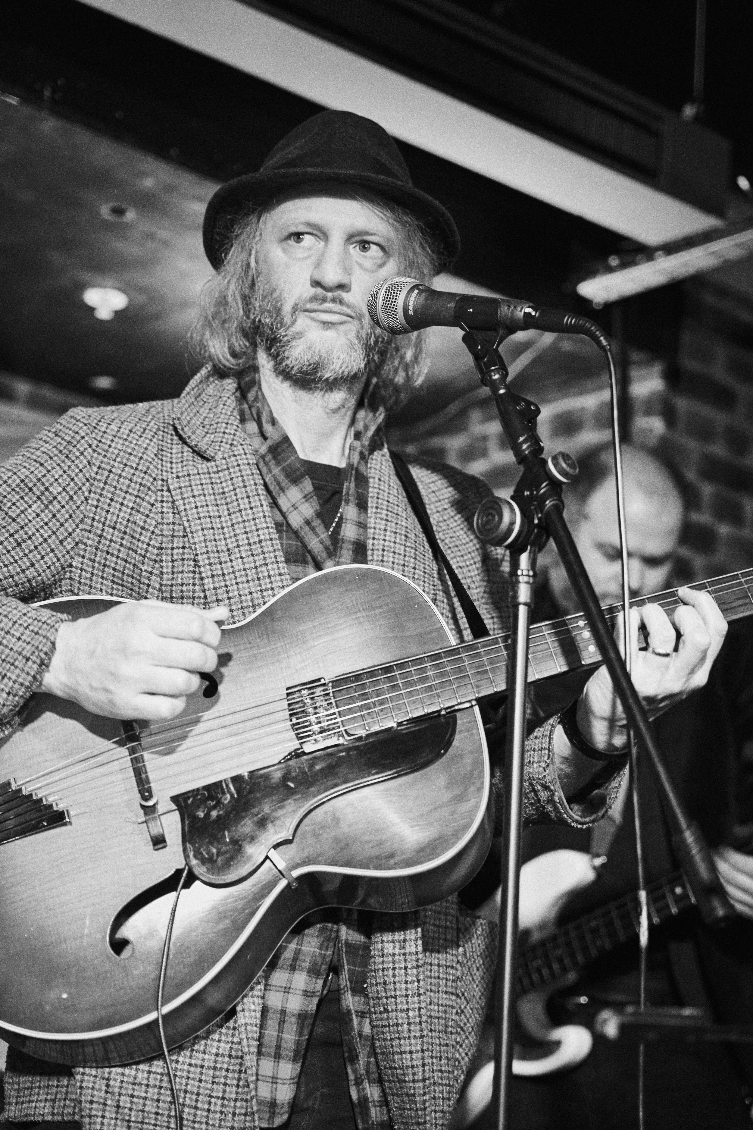 Black and white photo of a man wearing a hat, singing into a microphone, and playing an acoustic guitar on stage.