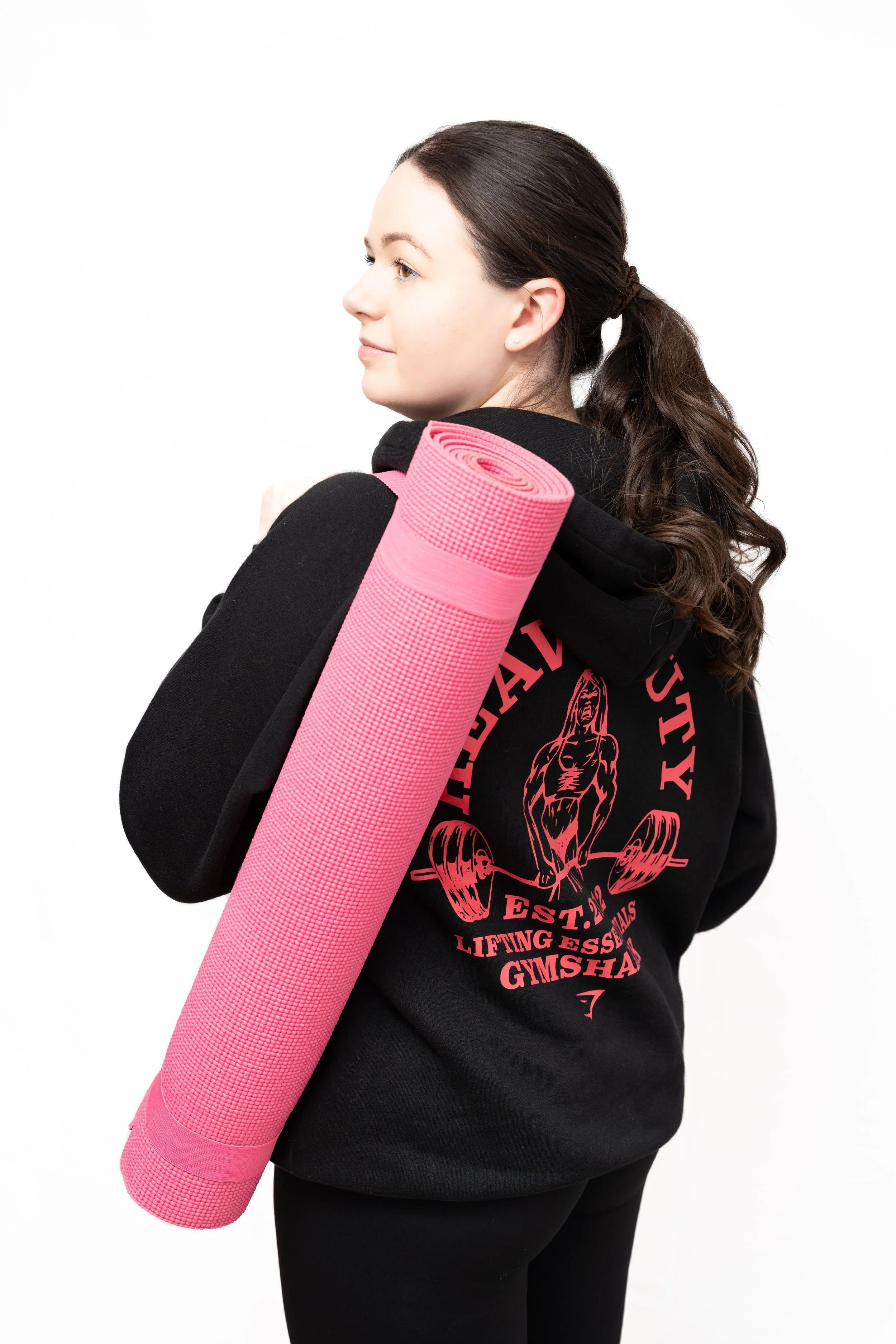 Young woman with long brown hair tied back, wearing a black sweatshirt with pink graphic design and text, carrying a pink yoga mat on her shoulder, standing against a plain white background.