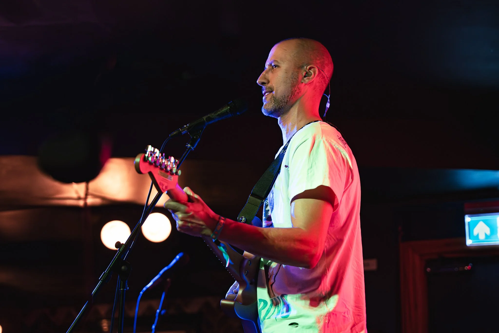 A male musician with a shaved head and beard performing on stage, singing into a microphone and holding a guitar, under colorful stage lights.