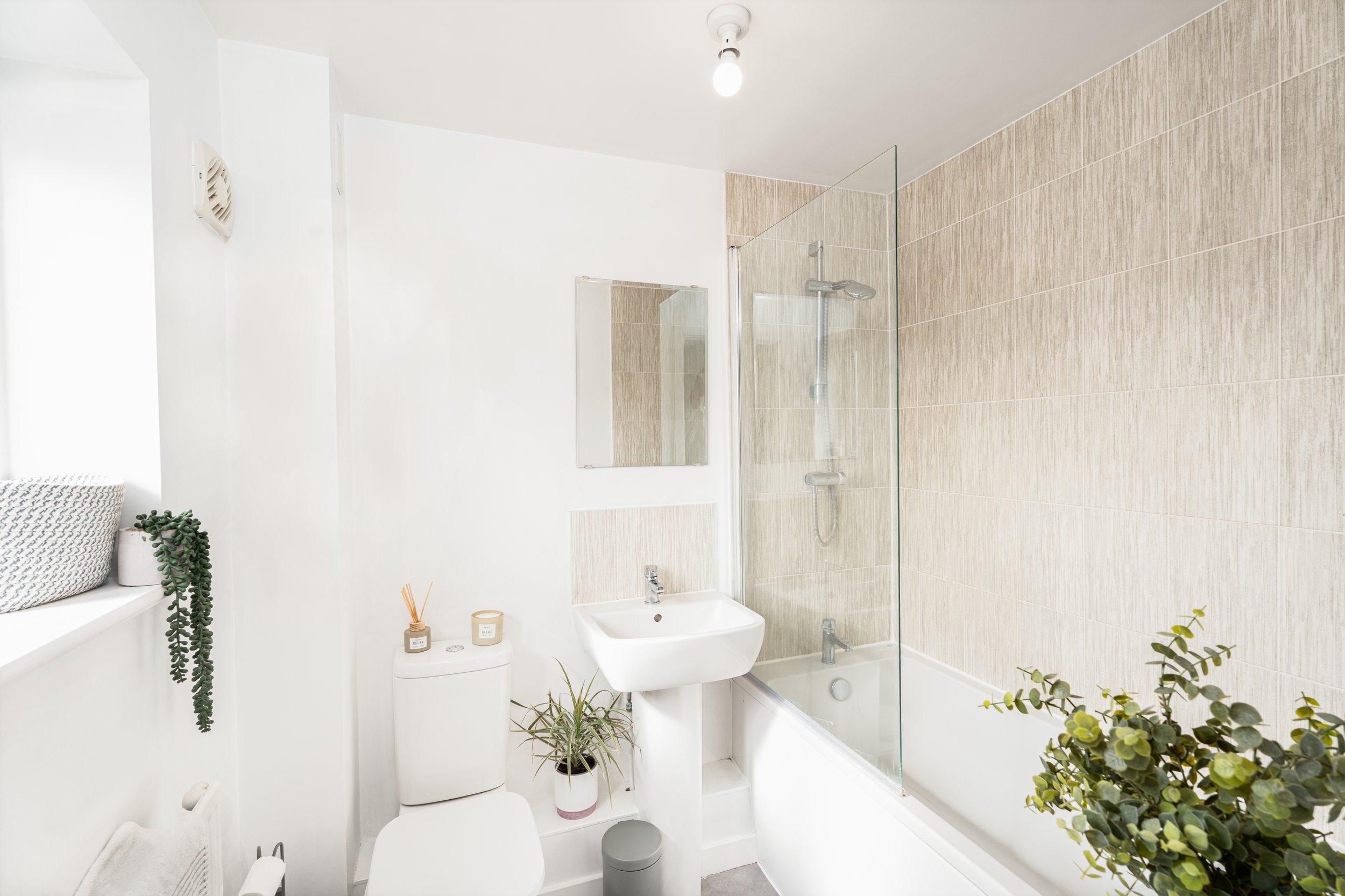 A modern, minimalist bathroom featuring a white toilet, a small white sink, a large mirror, and a shower with a glass partition and beige tiled wall. Decor includes green plants, candles, and a small woven basket on the window sill.