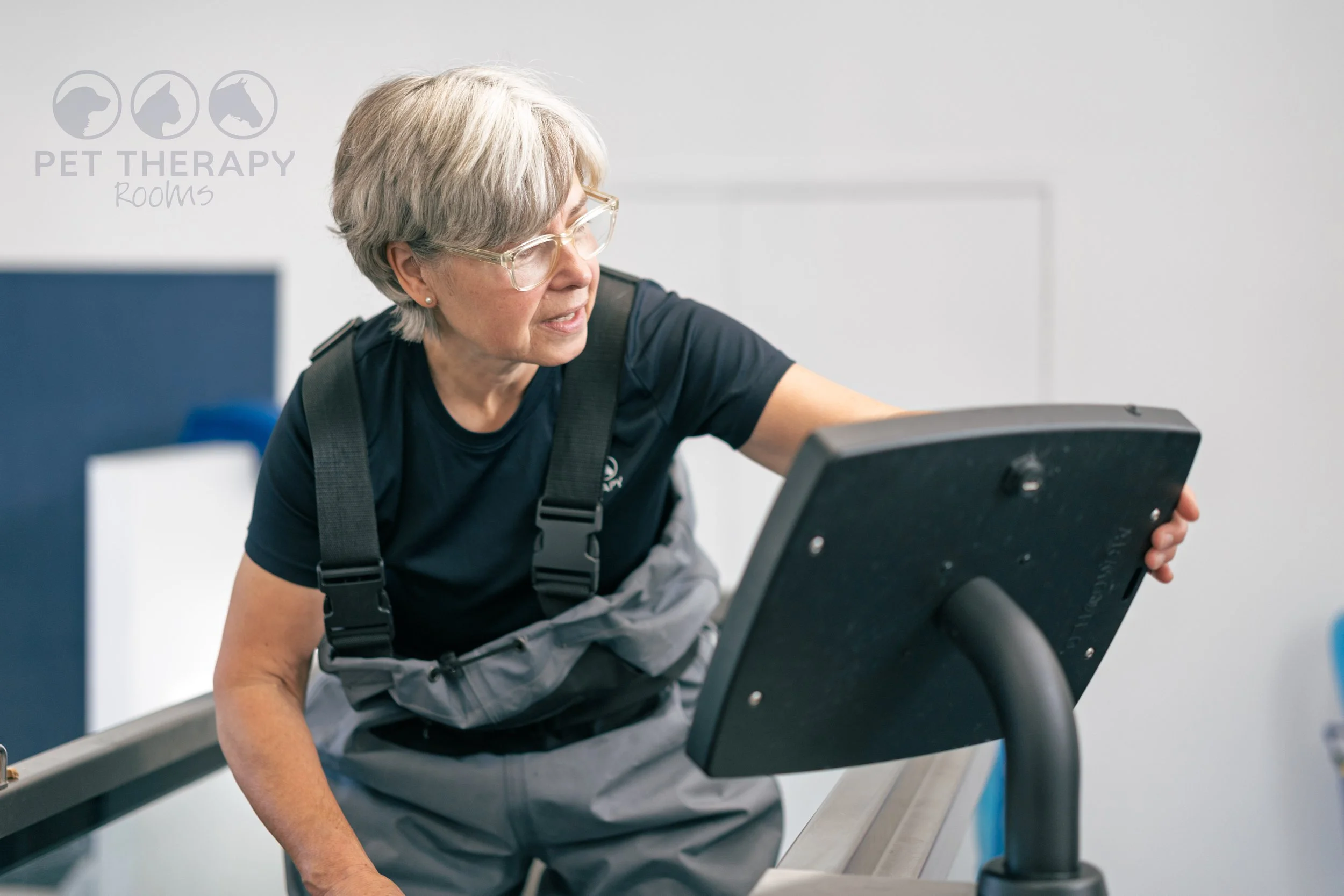 An woman with short grey hair and glasses operating a treadmill at a pet therapy center.