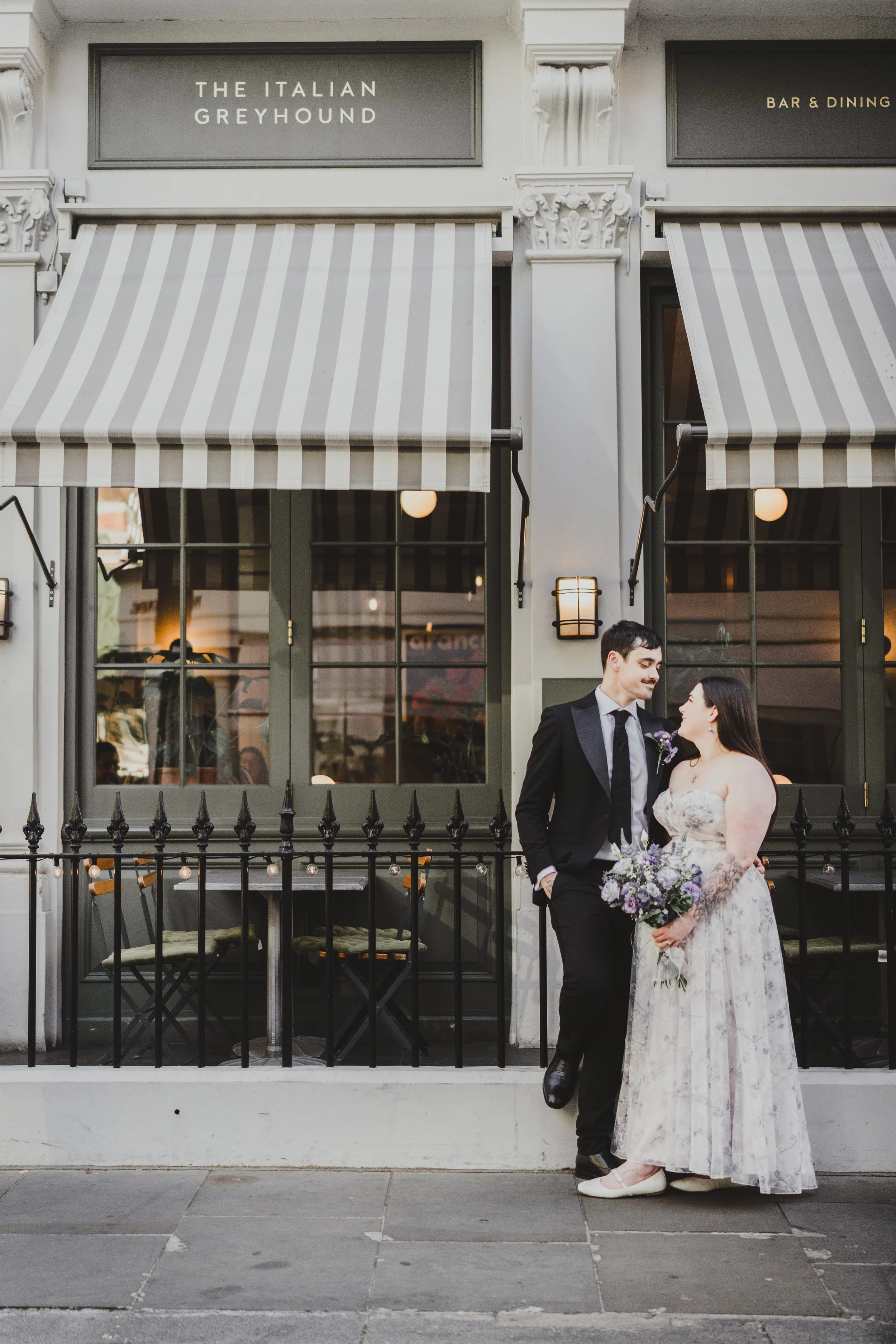 A bride and groom standing close together outside a restaurant called "The Italian Greyhound" during daytime. The groom is in a black tuxedo, and the bride is in a white lace wedding dress holding a bouquet. They are smiling and looking at each other.