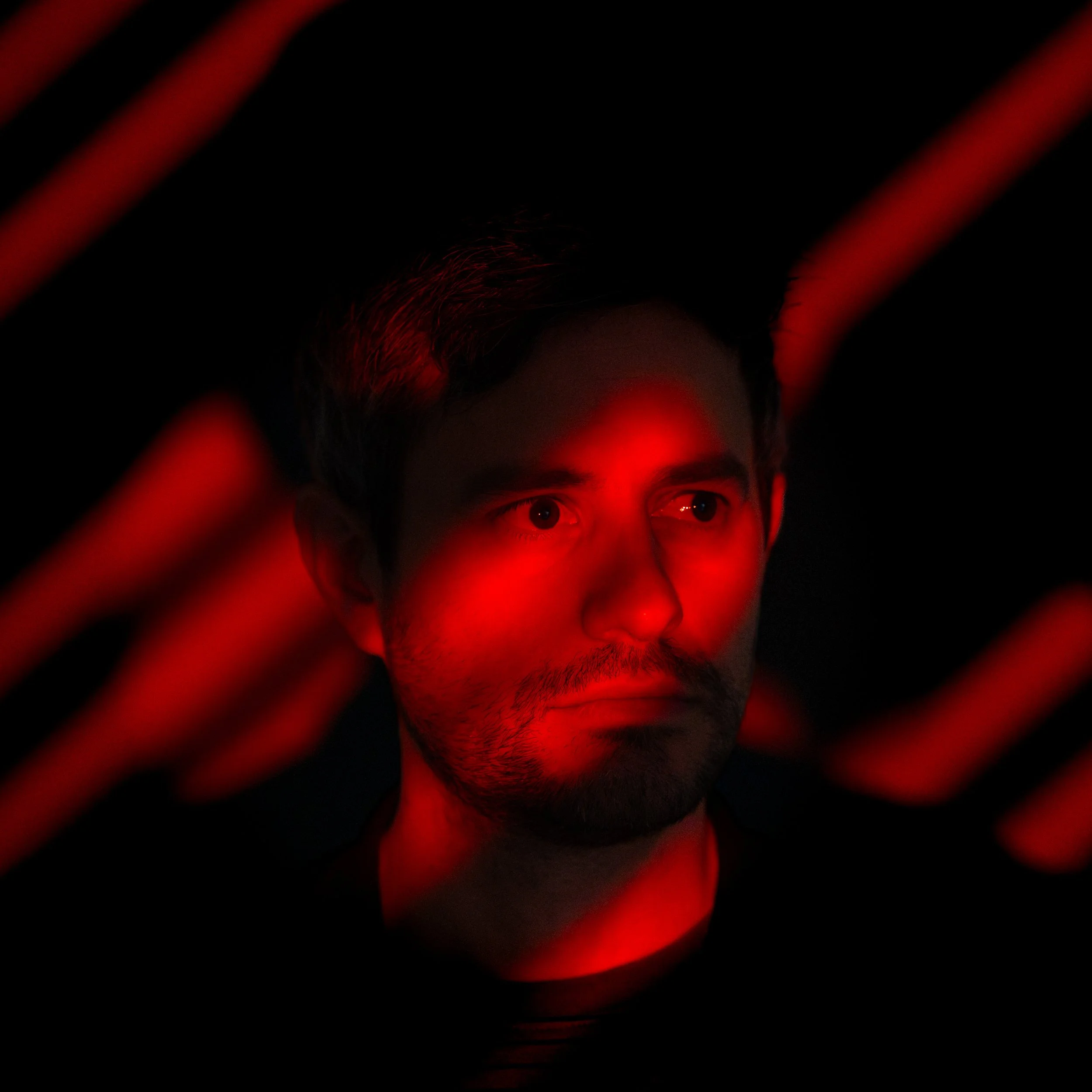 A person with dark hair and eyes gazes left under red lighting, with their face partially framed by dark, blurred red lines in the foreground.