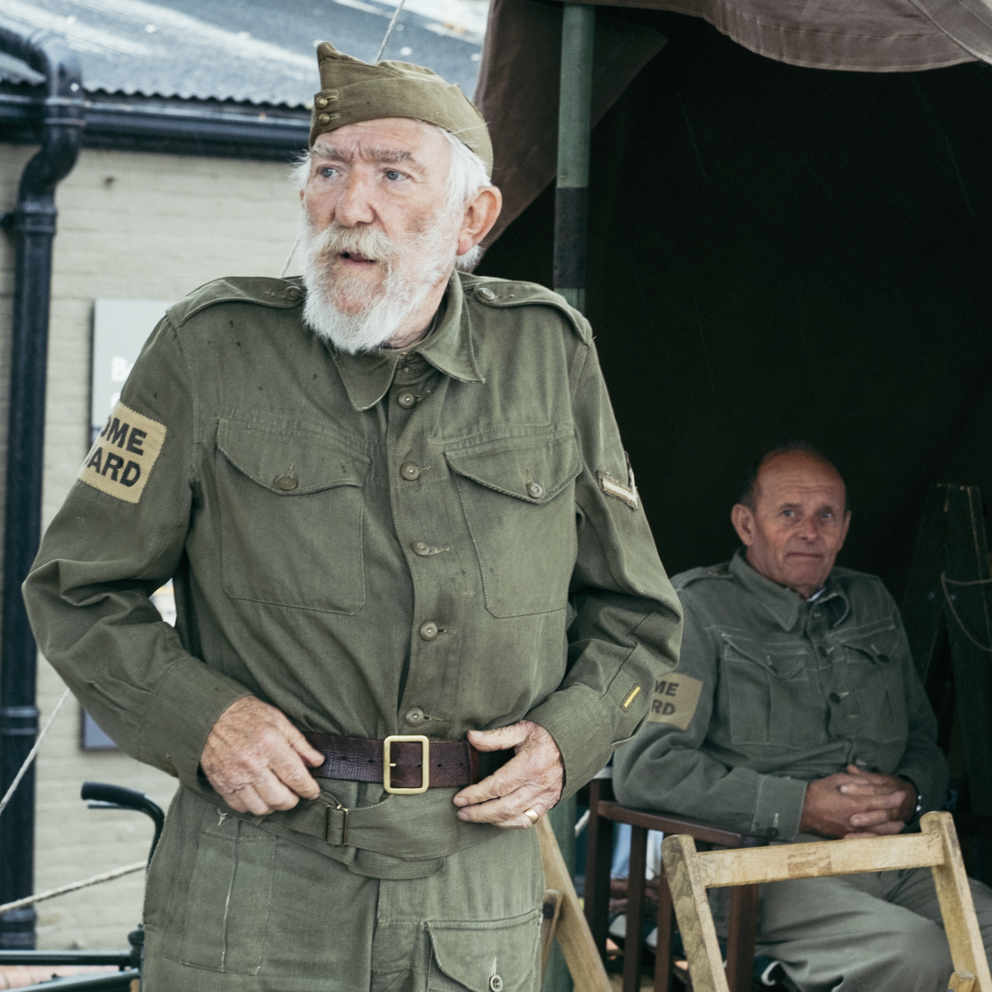 An elderly man dressed in a military uniform with a cap, standing outdoors. He has a white beard and grey hair. Another man is seated behind him, wearing a grey jacket, looking towards the camera.