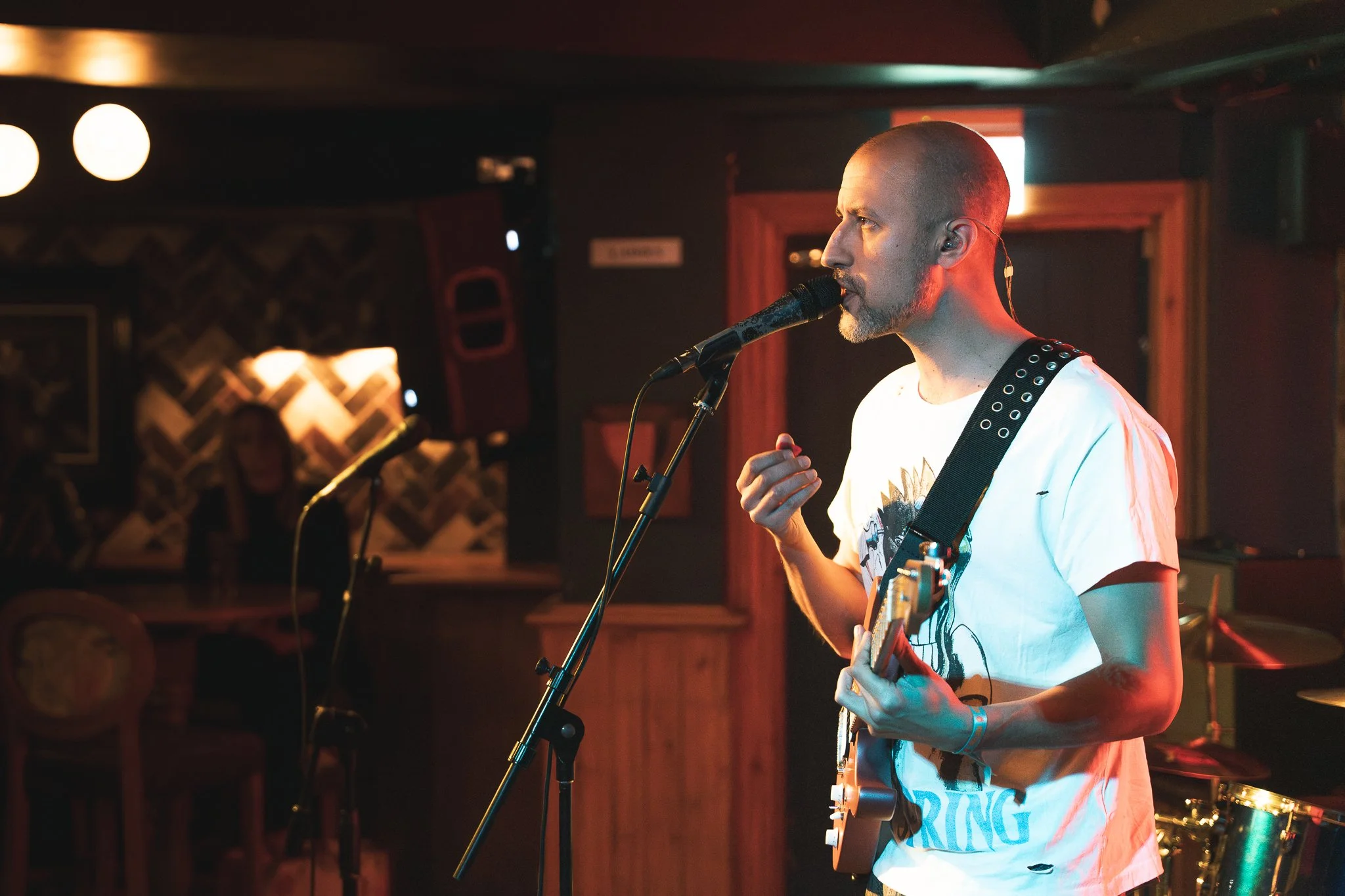 A man with a shaved head and beard performing on stage with a guitar and singing into a microphone in a dimly lit venue with some audience members in the background.