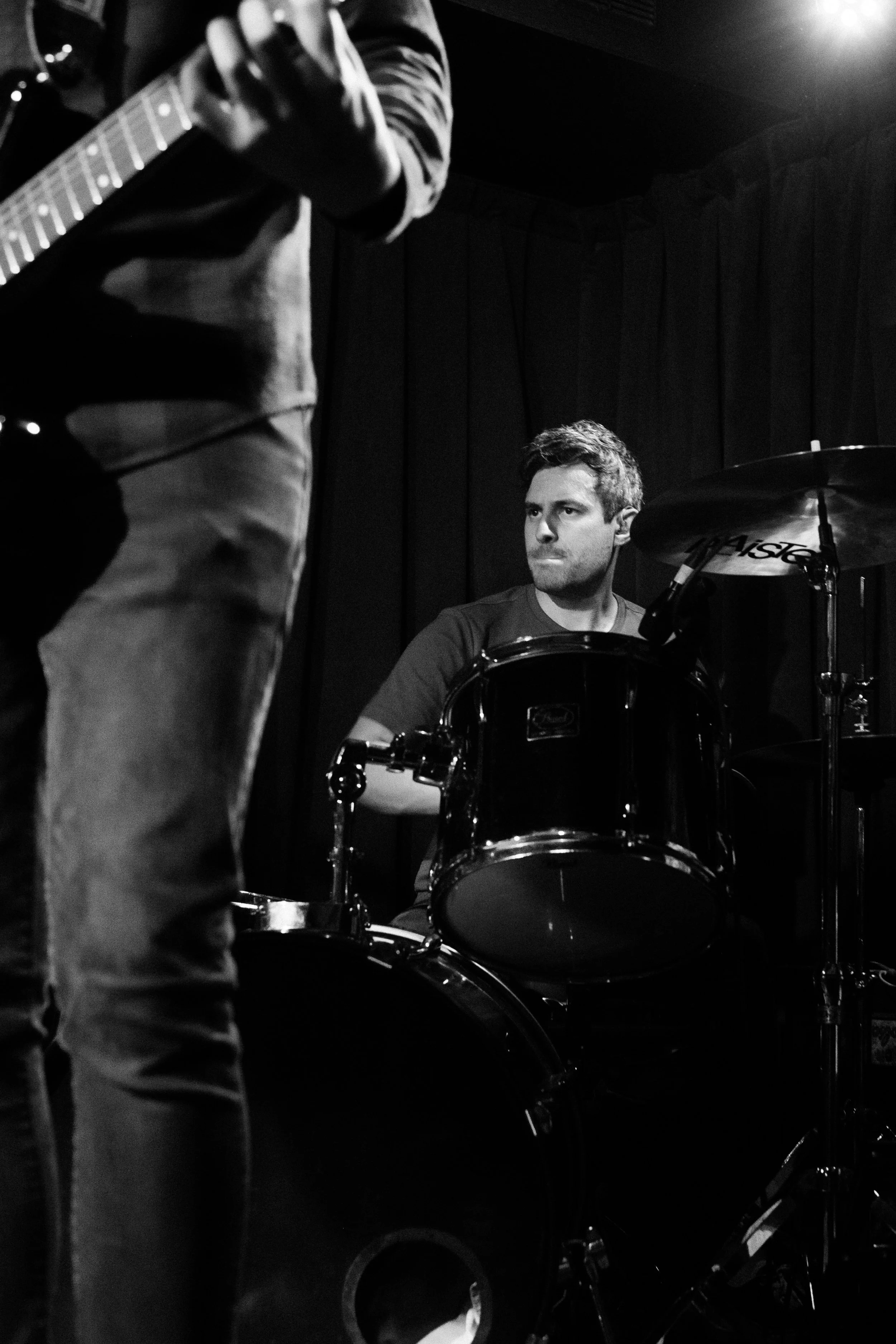 A man playing drums and another person playing guitar in a dimly lit music studio or performance space.