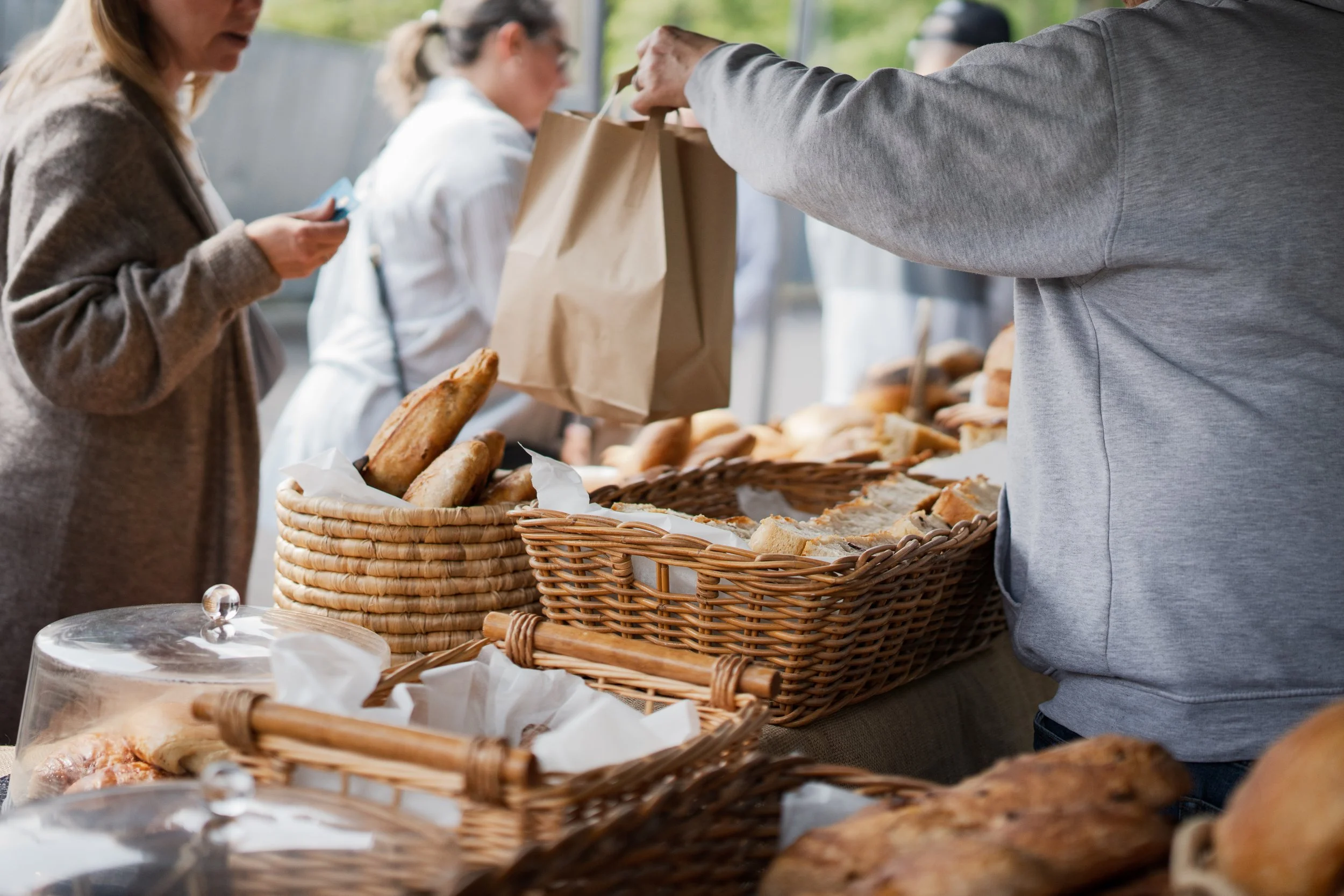 People selecting baked goods at an outdoor bakery stand with baskets of bread and pastries.