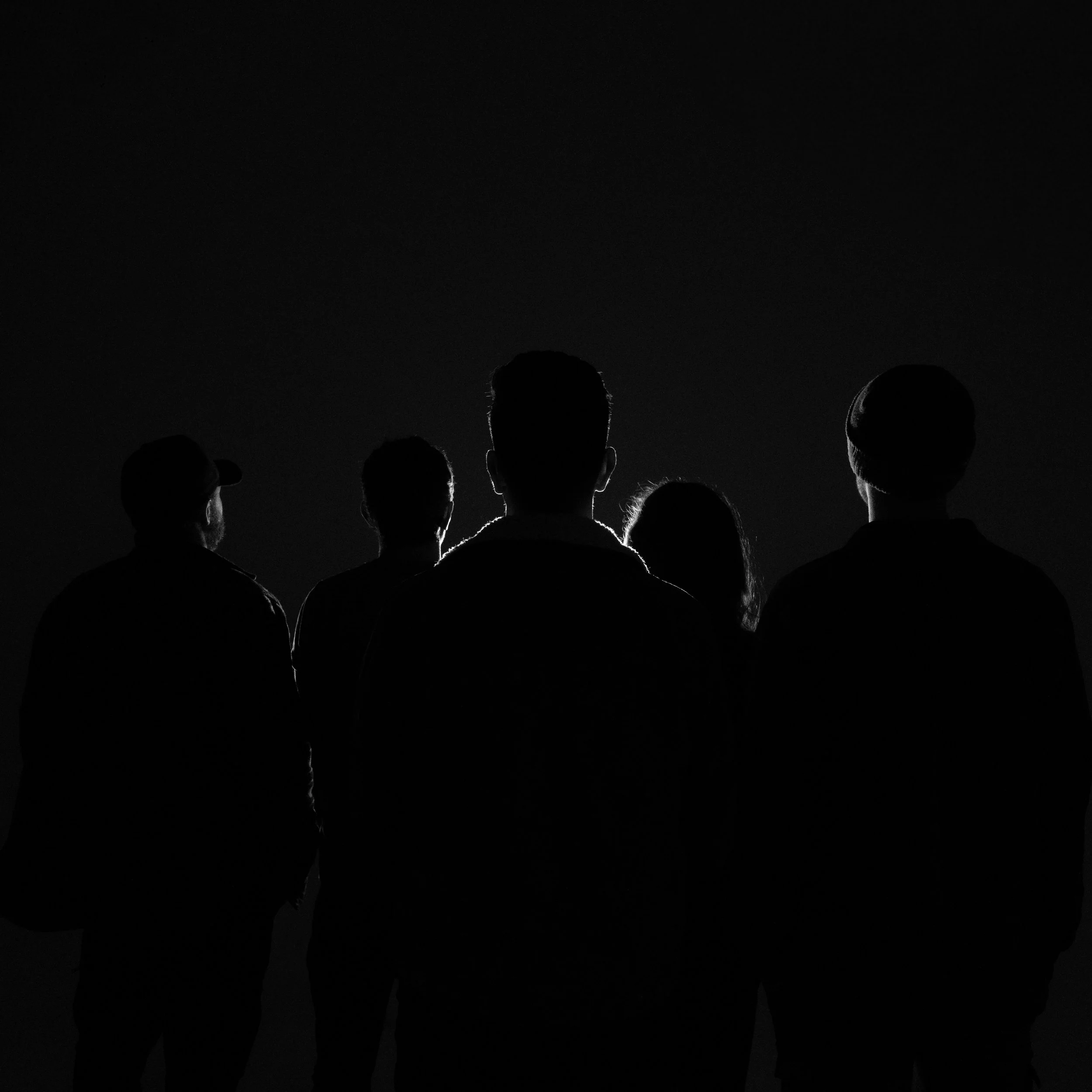 Silhouettes of several people standing together, their outline lit with a light on a black background. 