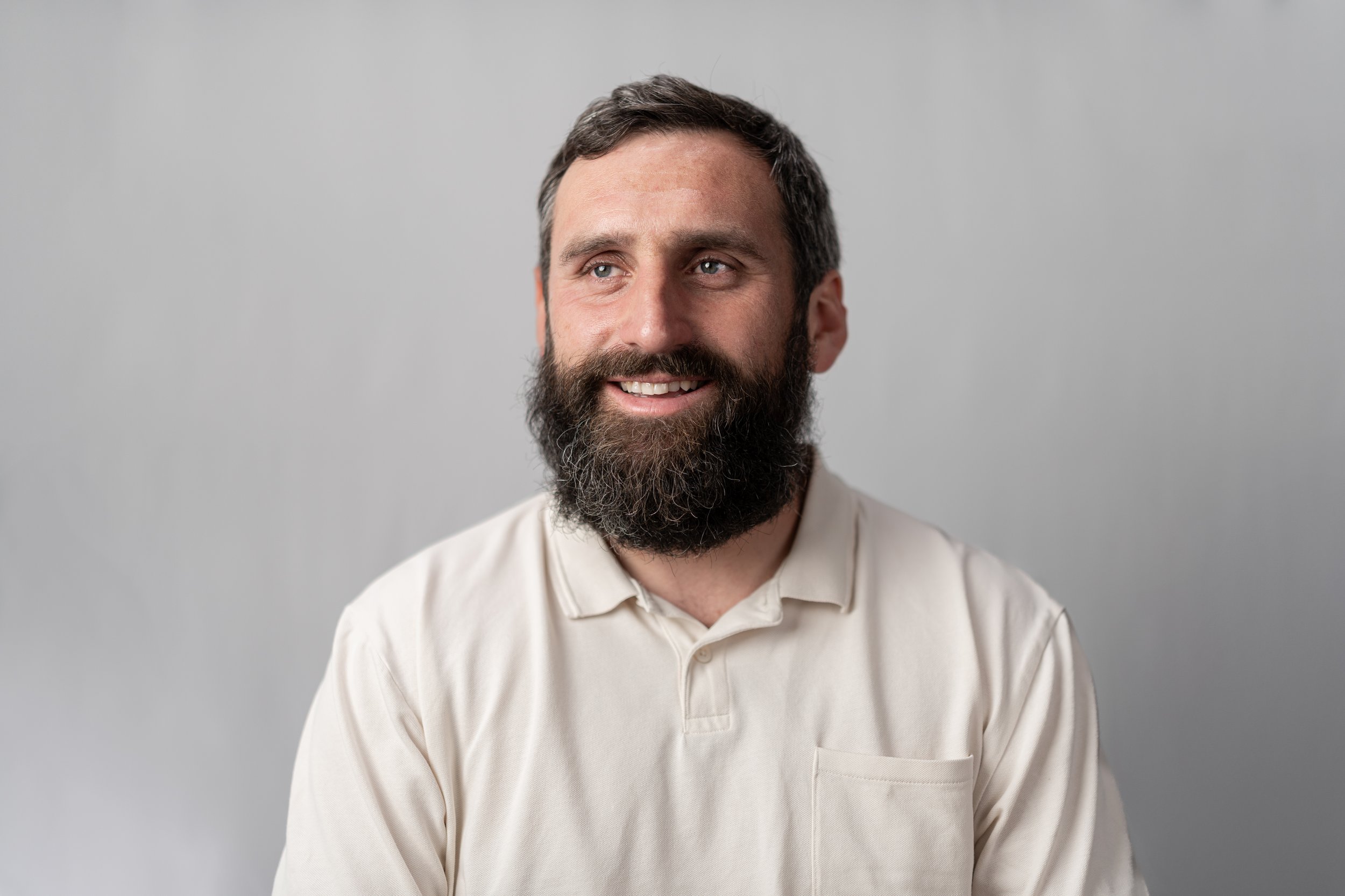 A man with a beard and short dark hair wearing a cream collared shirt, smiling slightly, against a plain gray background.