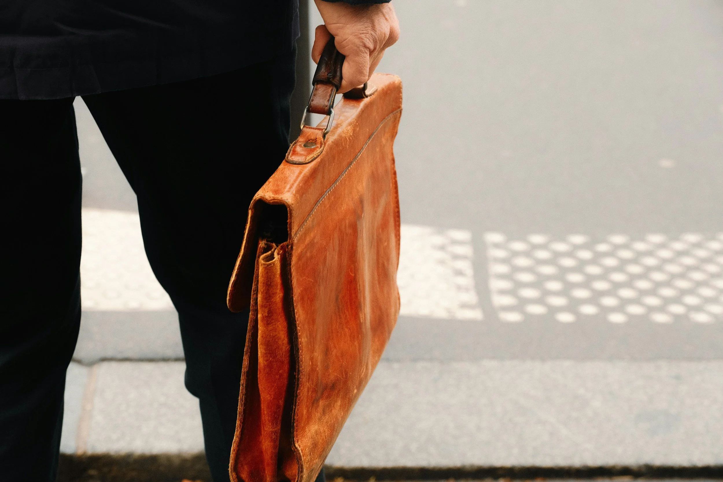 Person holding a brown leather briefcase in their right hand, wearing dark clothing, standing on a sidewalk with a vent or drain in the background.