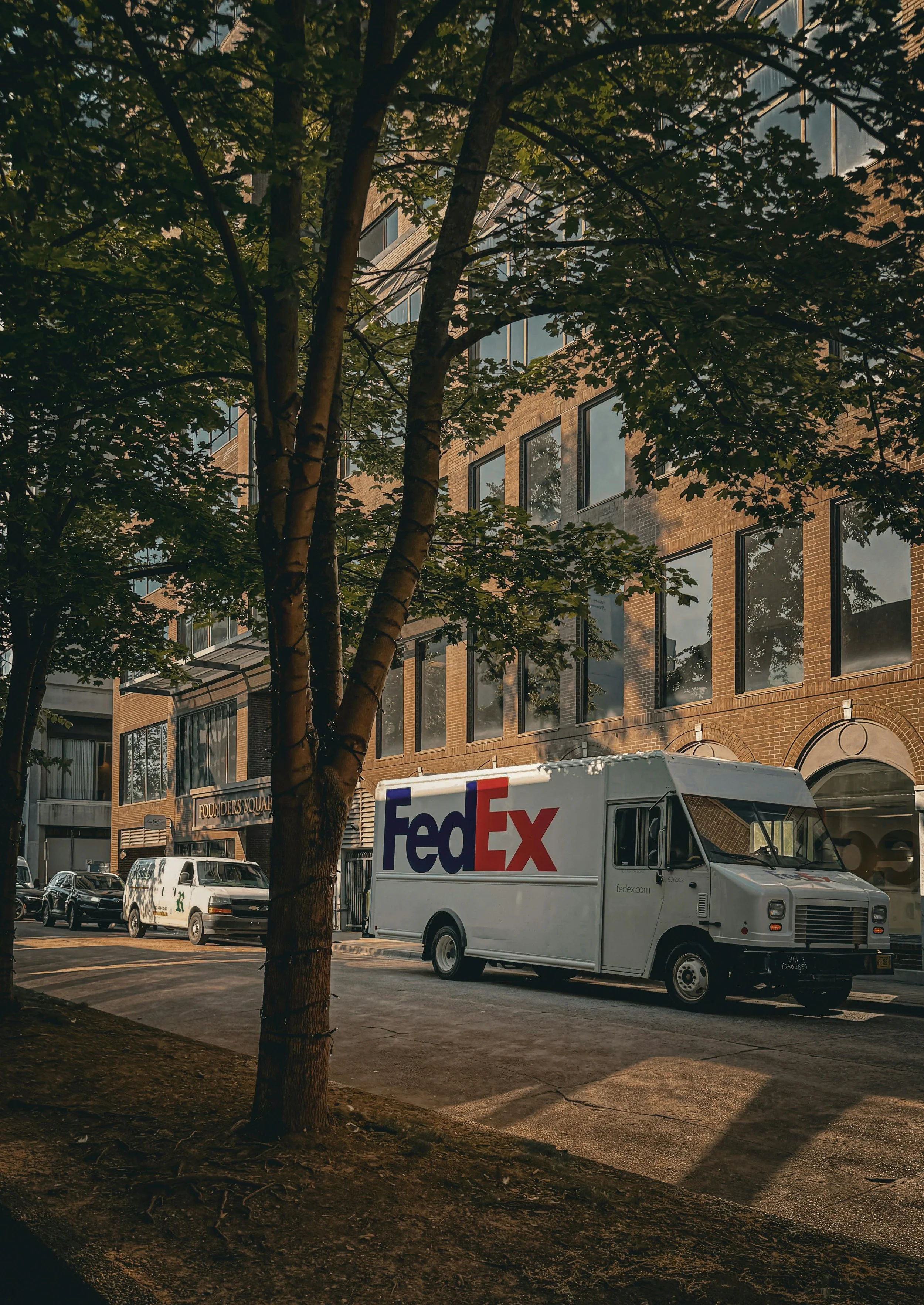 Street scene with a tree in the foreground, parked cars, including a FedEx delivery truck, and a brick building with large windows.