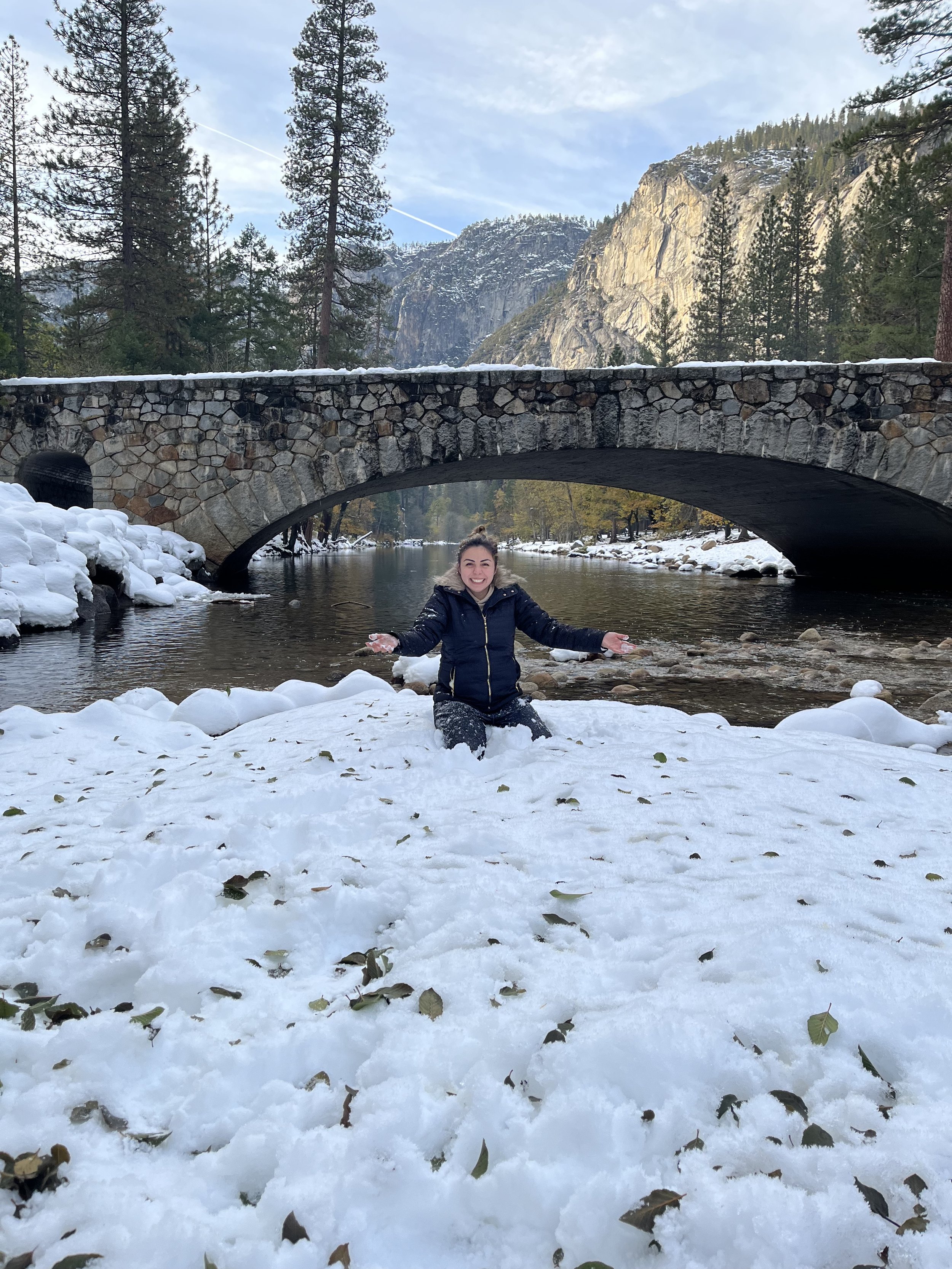 A woman kneeling in snow in front of a river with a stone bridge, surrounded by tall pine trees and mountains.