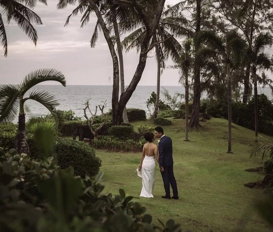 A couple in wedding attire, the woman in a white dress and the man in a dark suit, holding hands and walking in a lush garden near the ocean with palm trees and cloudy sky.